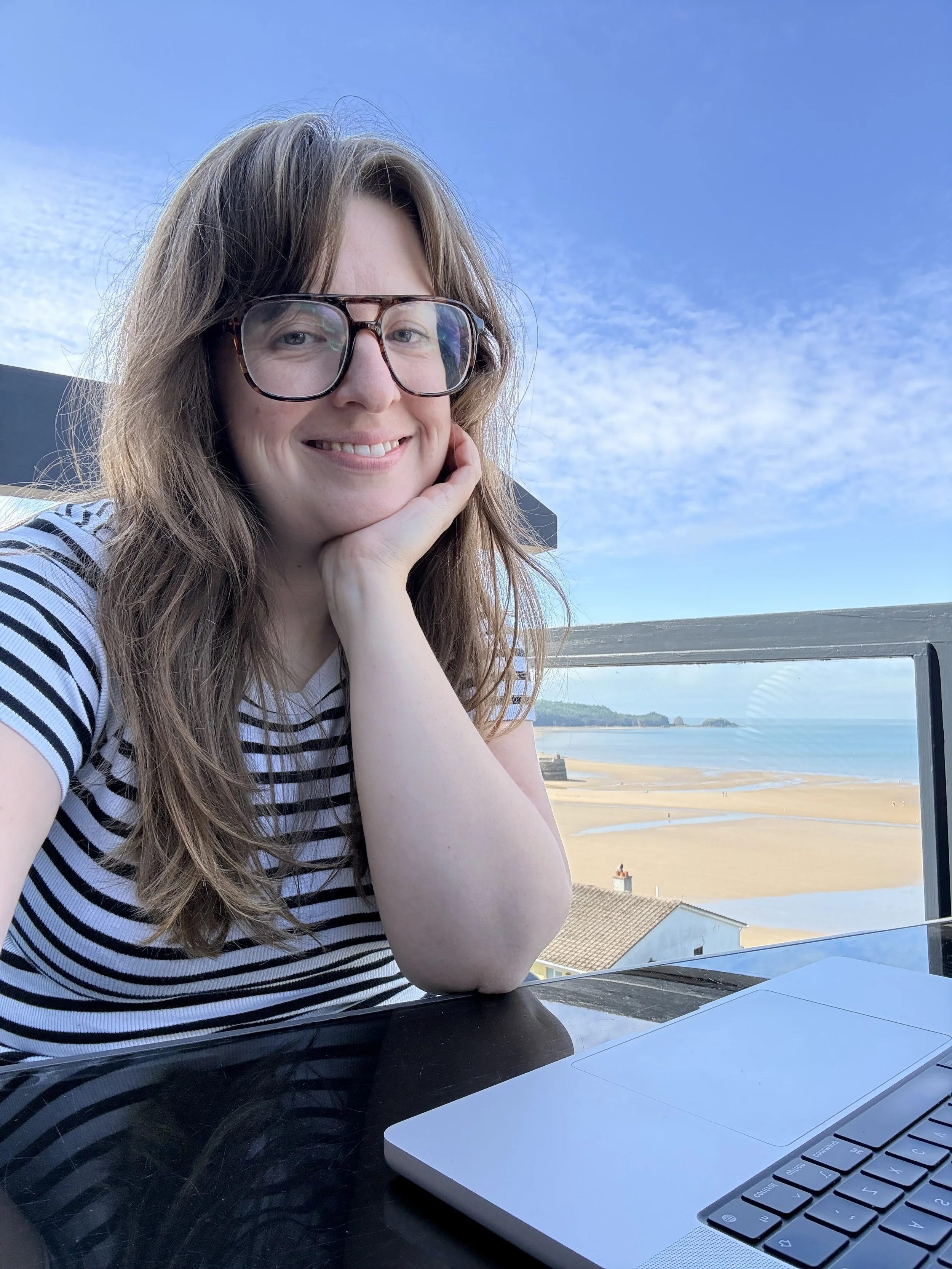A woman with long brown hair and glasses sitting outdoors at a table with a laptop, with a view of a beach and ocean in the background
