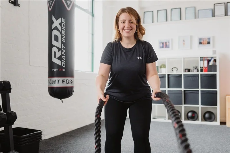 A smiling woman in workout clothes holding battle ropes in a gym.