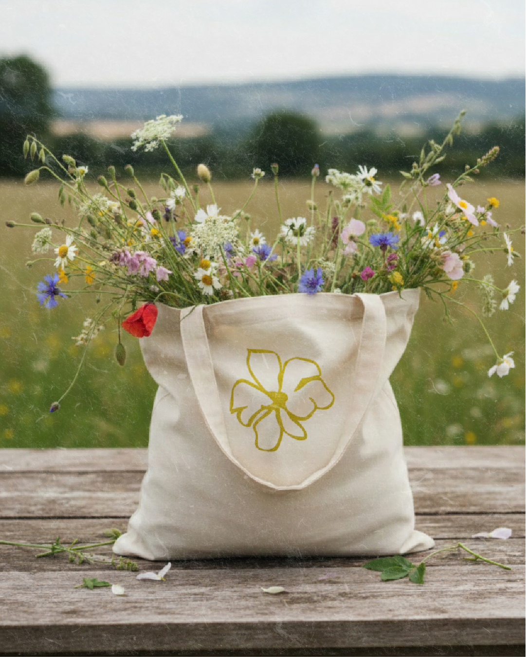 A white tote bag with a yellow outline of a flower on it, filled with wildflowers, sitting on a wooden surface with a rural landscape in the background.