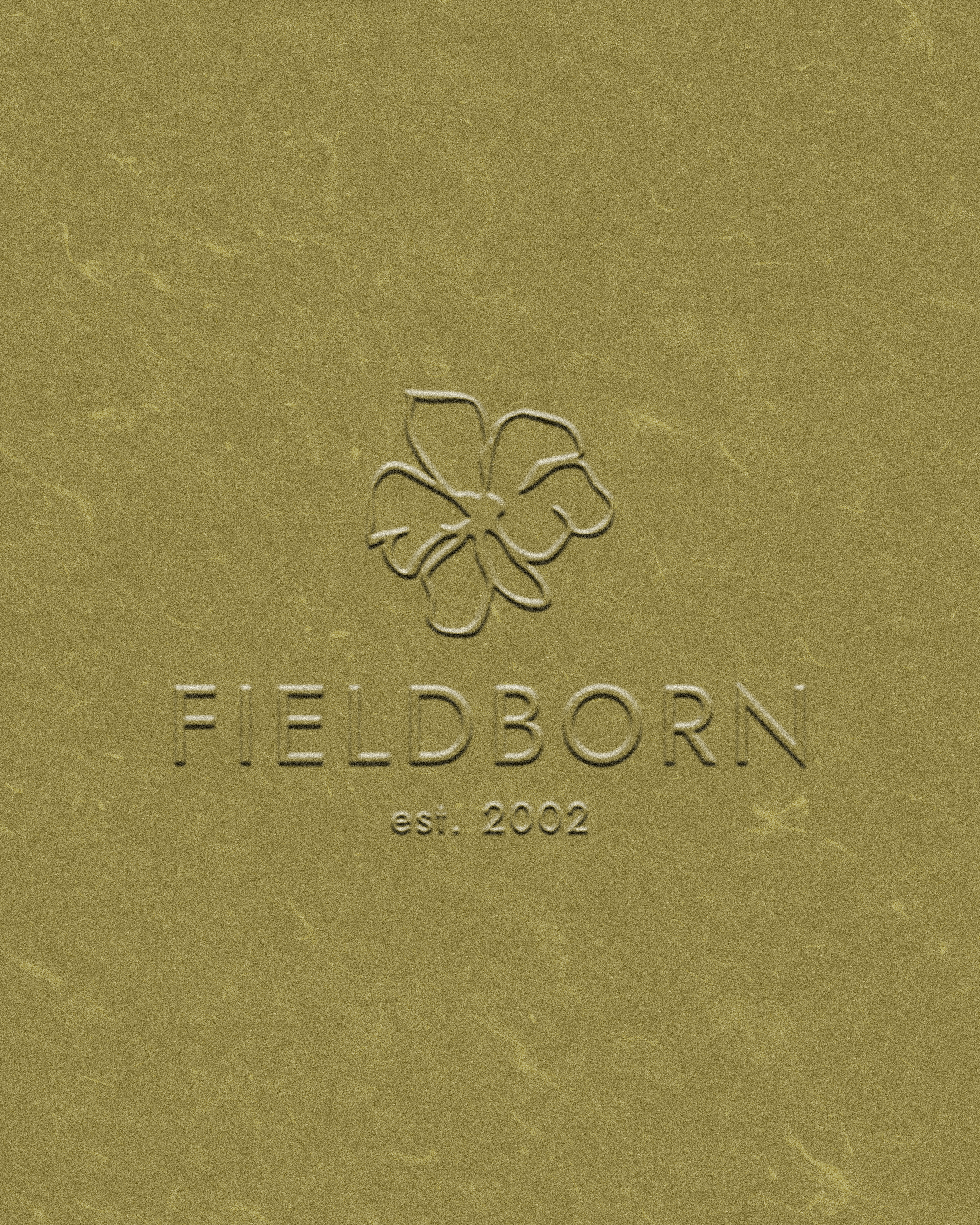 Close-up of a gold-colored surface with embossed text 'FIELDBORN est. 2002' and a flower outline logo above the text.
