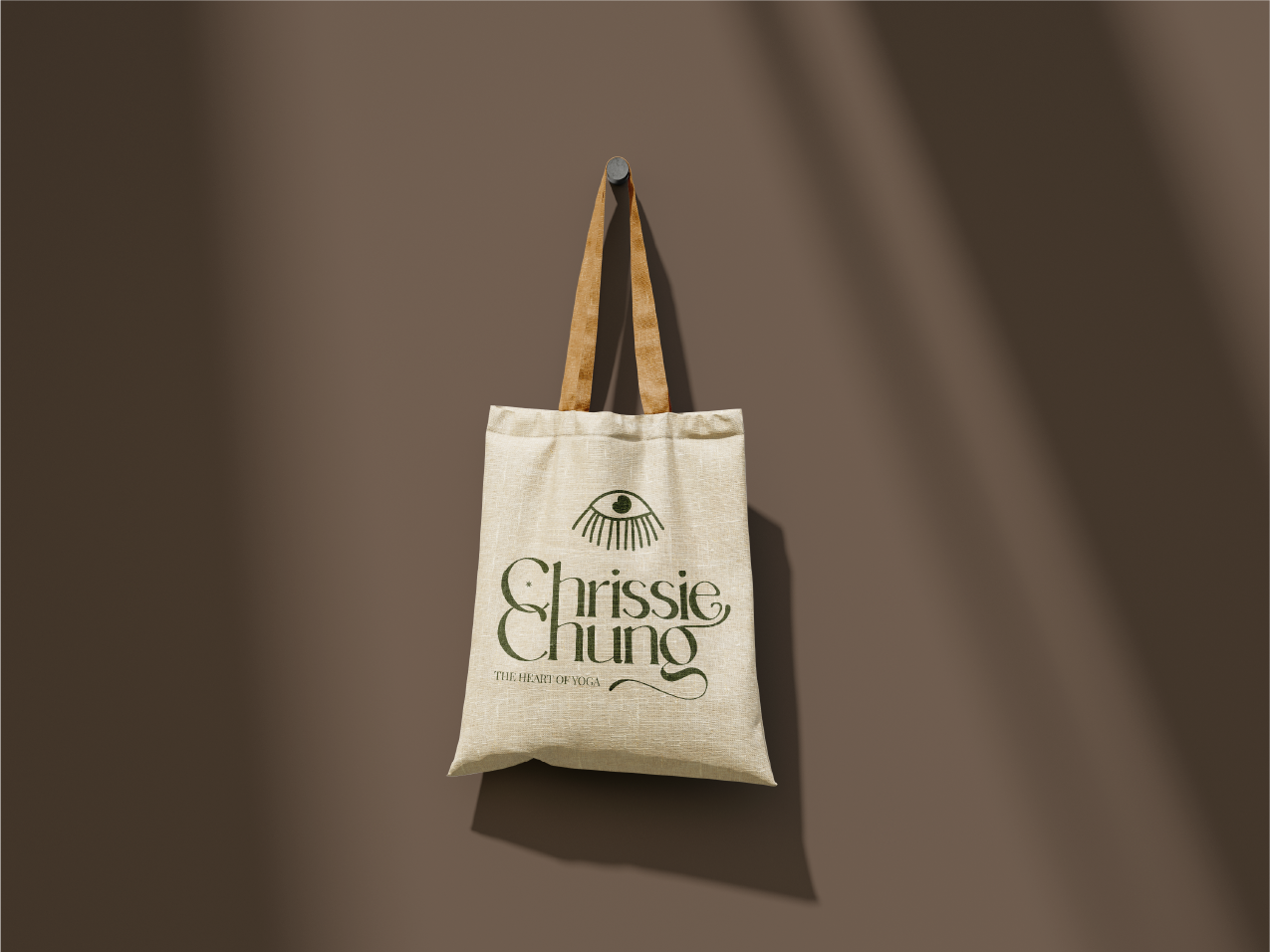 A beige tote bag with brown handles hanging on a wall hook, with the text "Chrissie Chung The Heart of Yoga" and a stylized eye logo printed on the front.