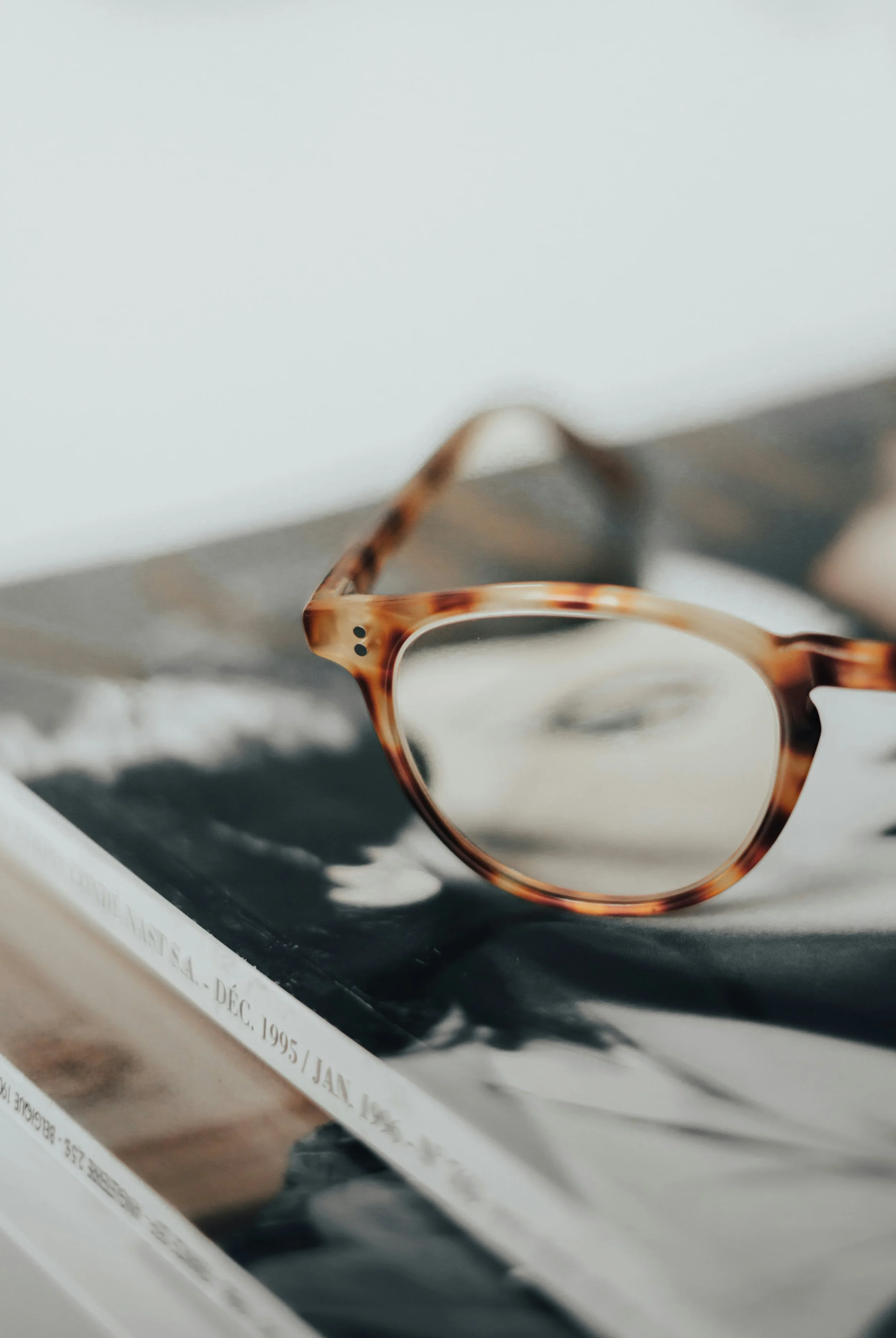 Close-up of a pair of tortoiseshell eyeglasses resting on a black surface, with a blurred piece of paper or magazine underneath.