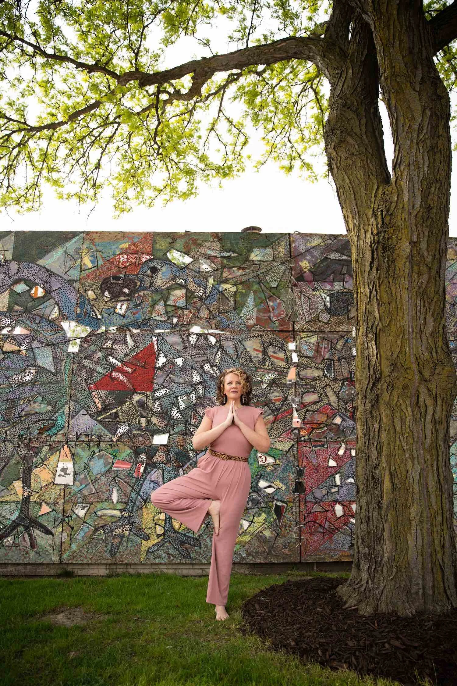 A woman practicing yoga outdoors in a tree pose, standing on one leg with hands pressed together in front of her chest, in front of a colorful mosaic wall and a large tree.