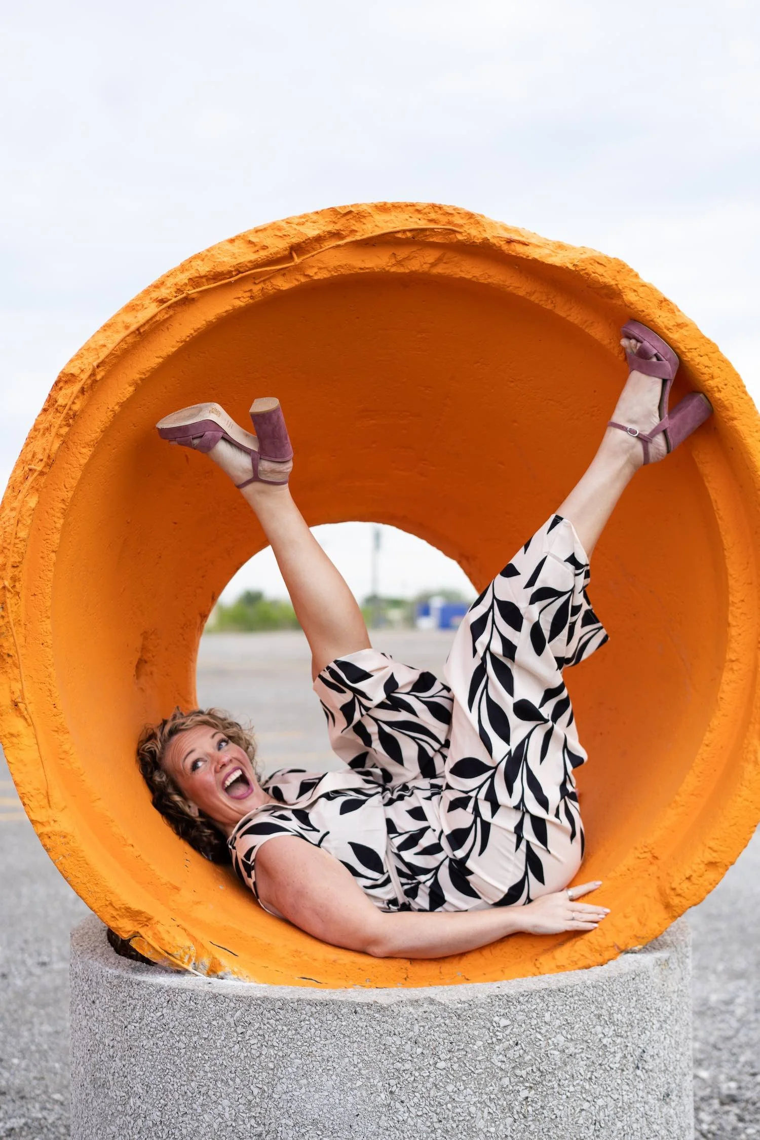 Woman lying inside a large, hollow, orange concrete pipe with her legs in the air, smiling and laughing.
