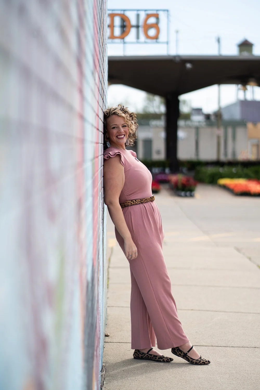 A woman with curly blonde hair, wearing a pink jumpsuit with ruffled sleeves and leopard print shoes, leaning against a colorful mural wall, smiling at the camera.