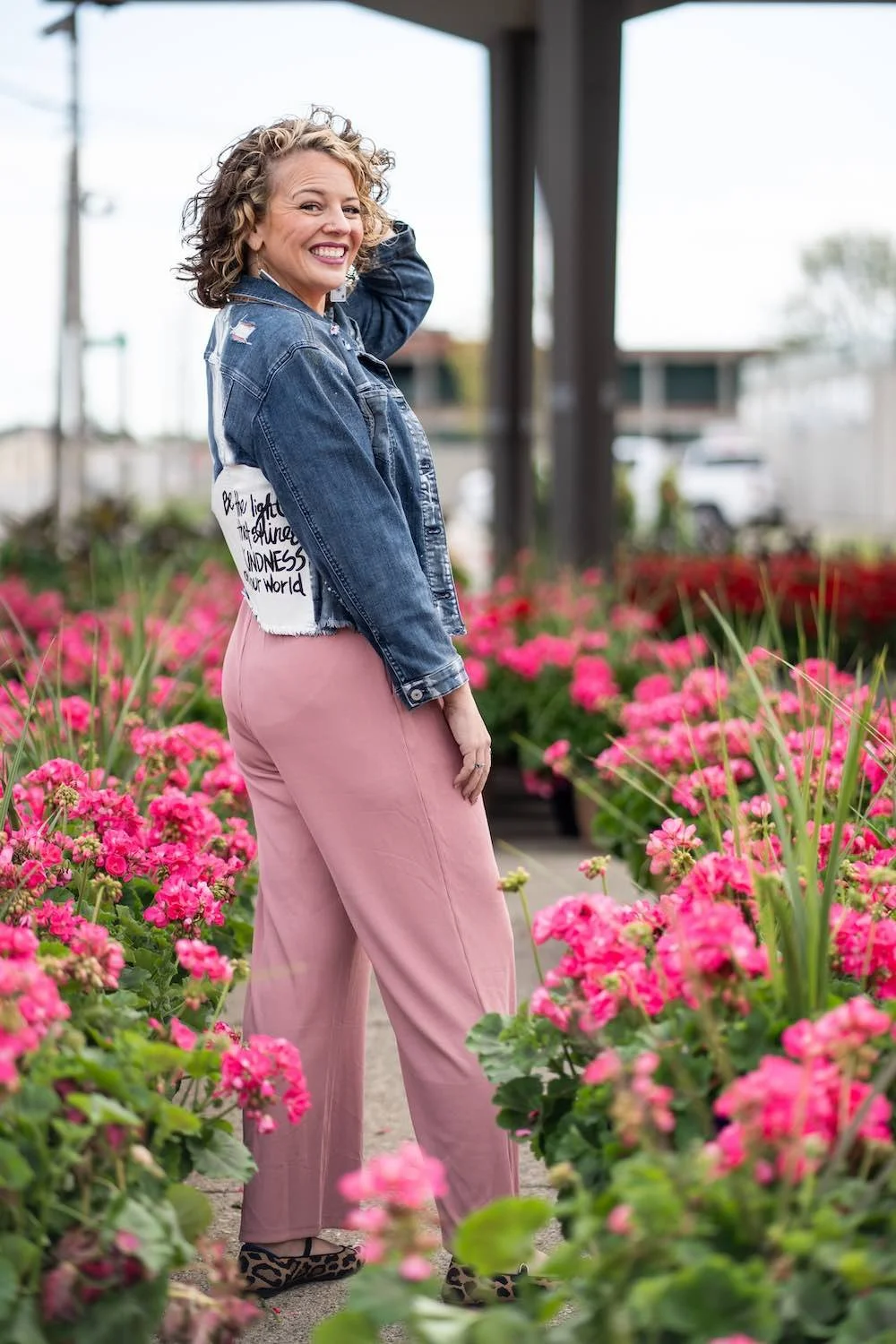 A woman with curly blonde hair, wearing a denim jacket with a patch on the back that reads 'Be the light that shines in kindness of our world,' pink pants, and leopard print shoes, standing in a garden of pink flowers and smiling at the camera.