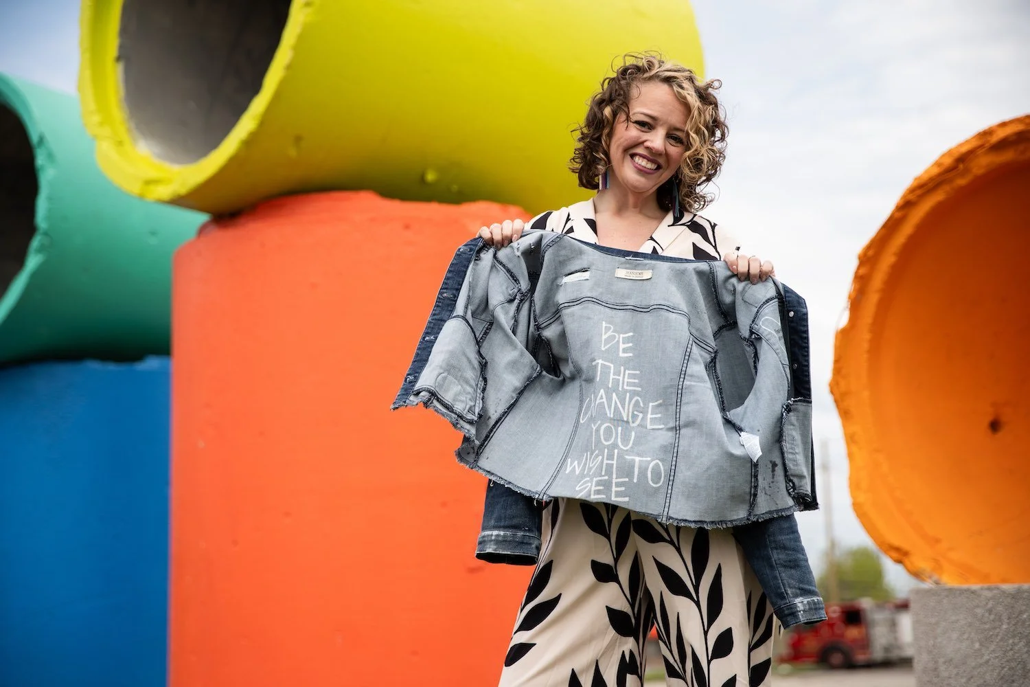 Woman smiling and holding a denim jacket with embroidered text, standing outdoors among large colorful pipe sculptures.