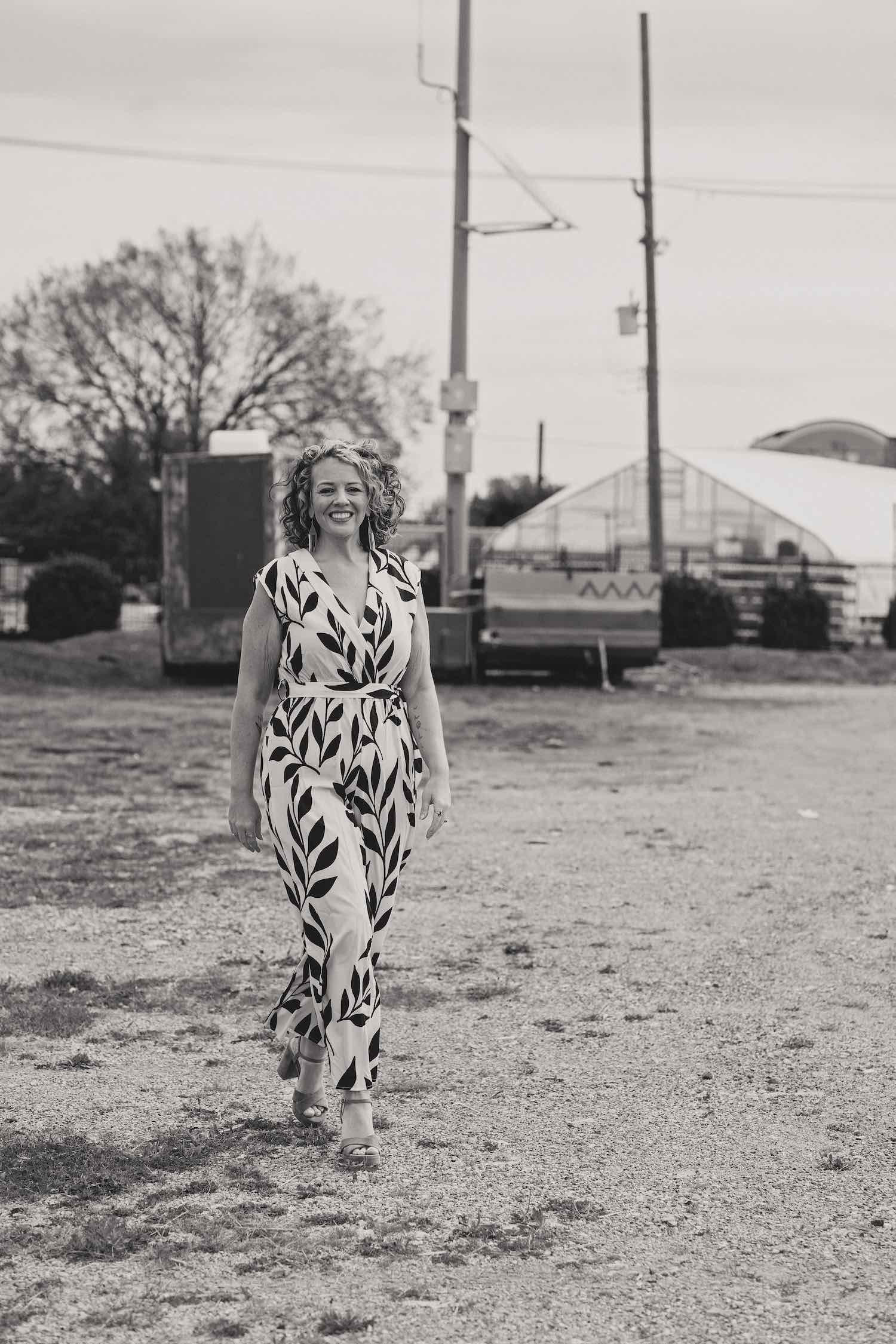 A woman with curly hair smiling, walking on a gravel path outdoors, wearing a patterned jumpsuit and high heels, with trees, structures, and power poles in the background.
