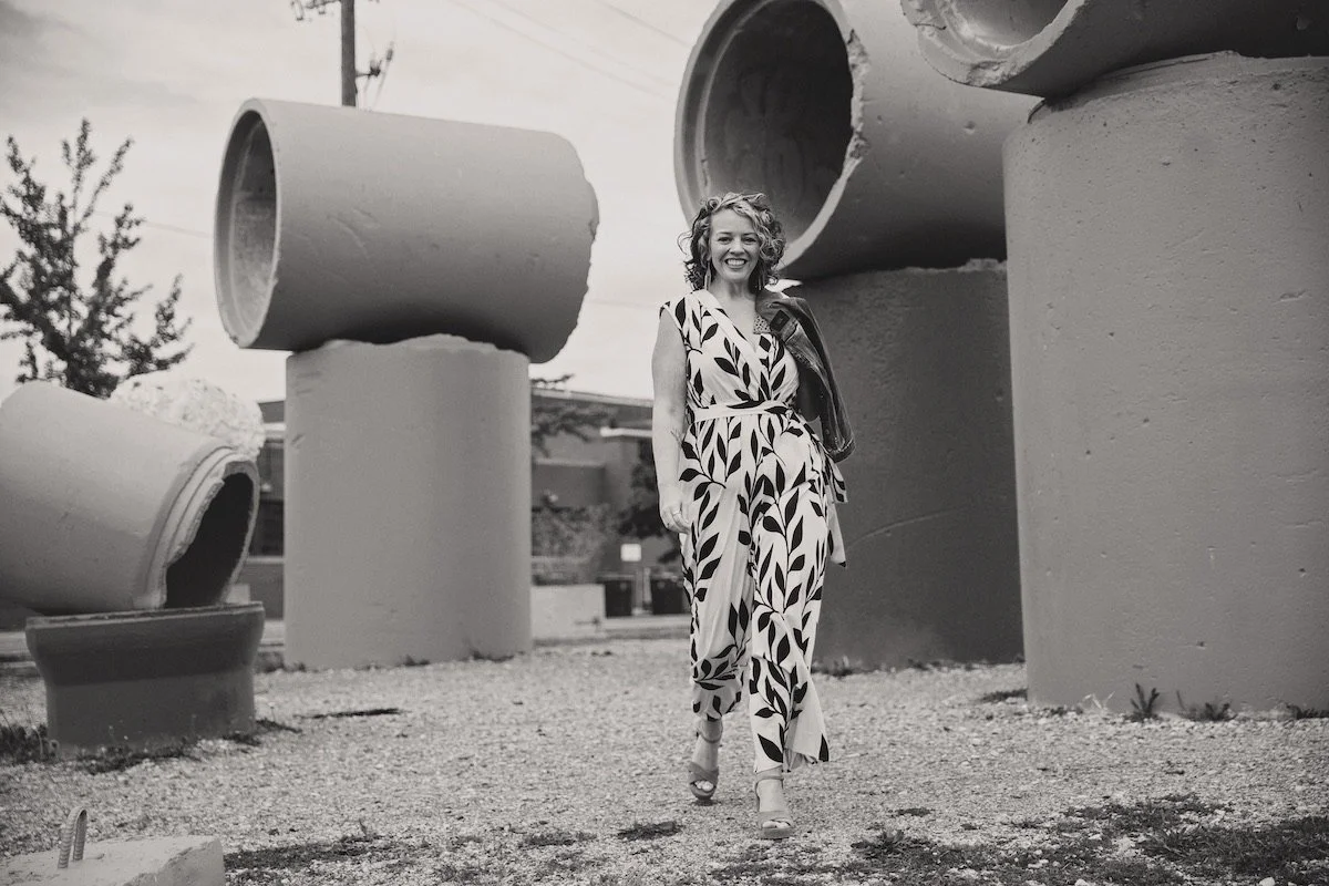 A woman walking outdoors among large cylindrical concrete pipes, smiling and holding a jacket over her shoulder in a black-and-white photo.