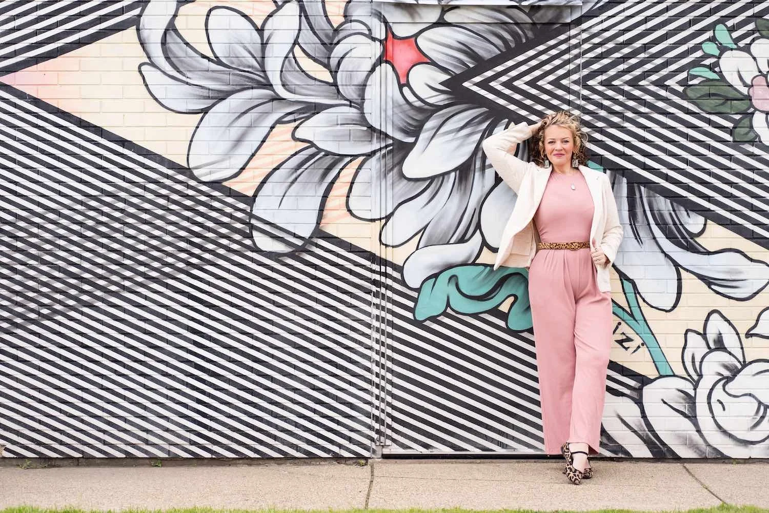 A woman in pink pants and a pink top, wearing a white blazer and leopard print heels, standing in front of a large mural of flowers on a brick wall, outdoors.