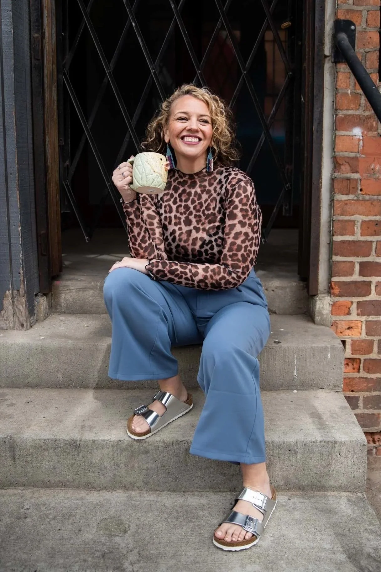 A woman sitting on concrete steps in front of a brick building, holding a mug resembling a cauliflower, smiling, wearing a leopard print top, blue pants, and sandals.