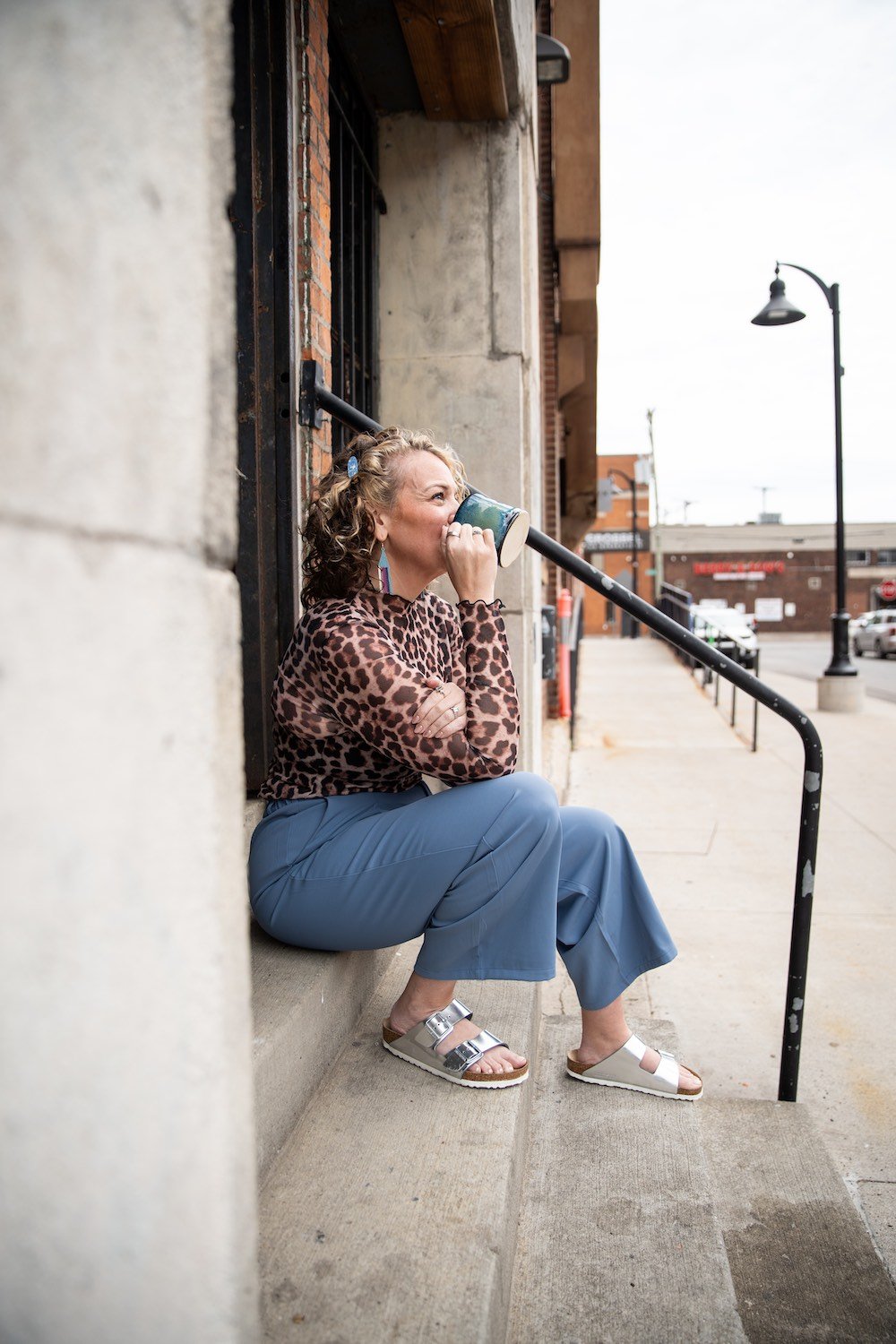 A woman with curly hair, wearing a leopard print top, sitting on a sidewalk in an urban area, drinking from a mug. She has earrings, a ring, and sandals, with buildings and streetlights in the background.