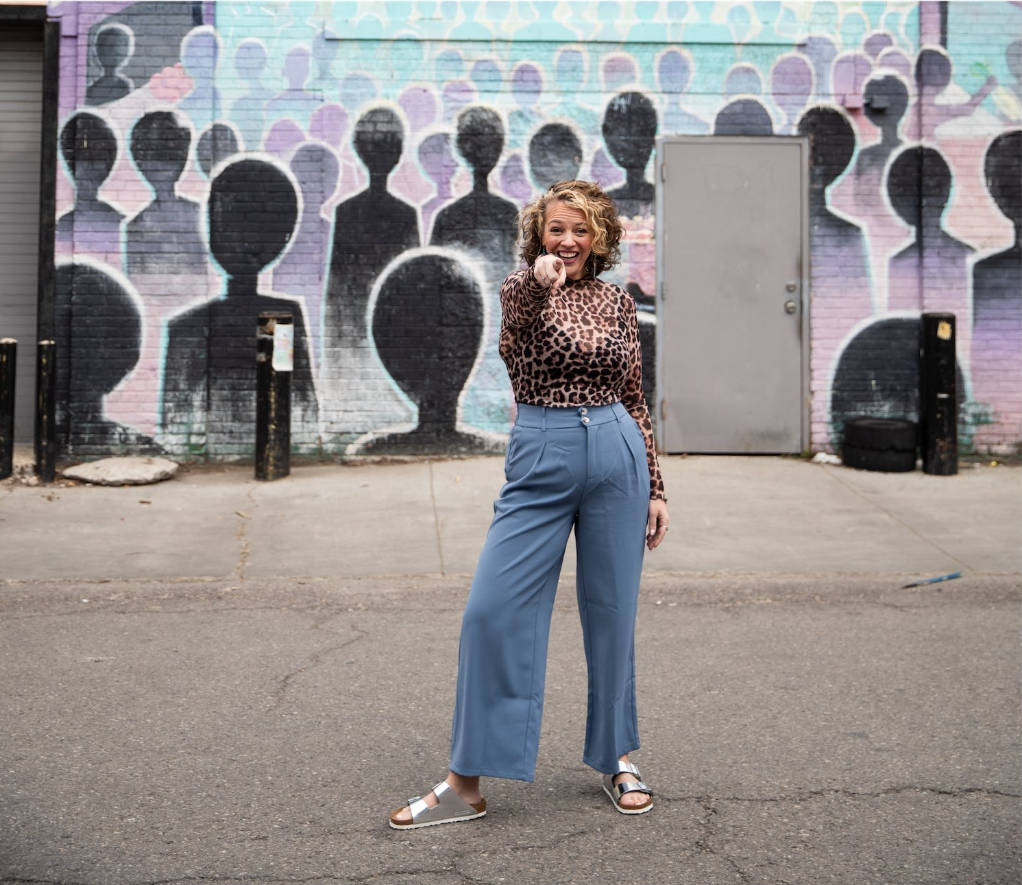 A woman with curly blonde hair, wearing a leopard print long-sleeve top and blue wide-leg pants, is standing on a city street pointing at the camera with a smile. Behind her is a vibrant mural of human silhouettes painted on a brick wall.