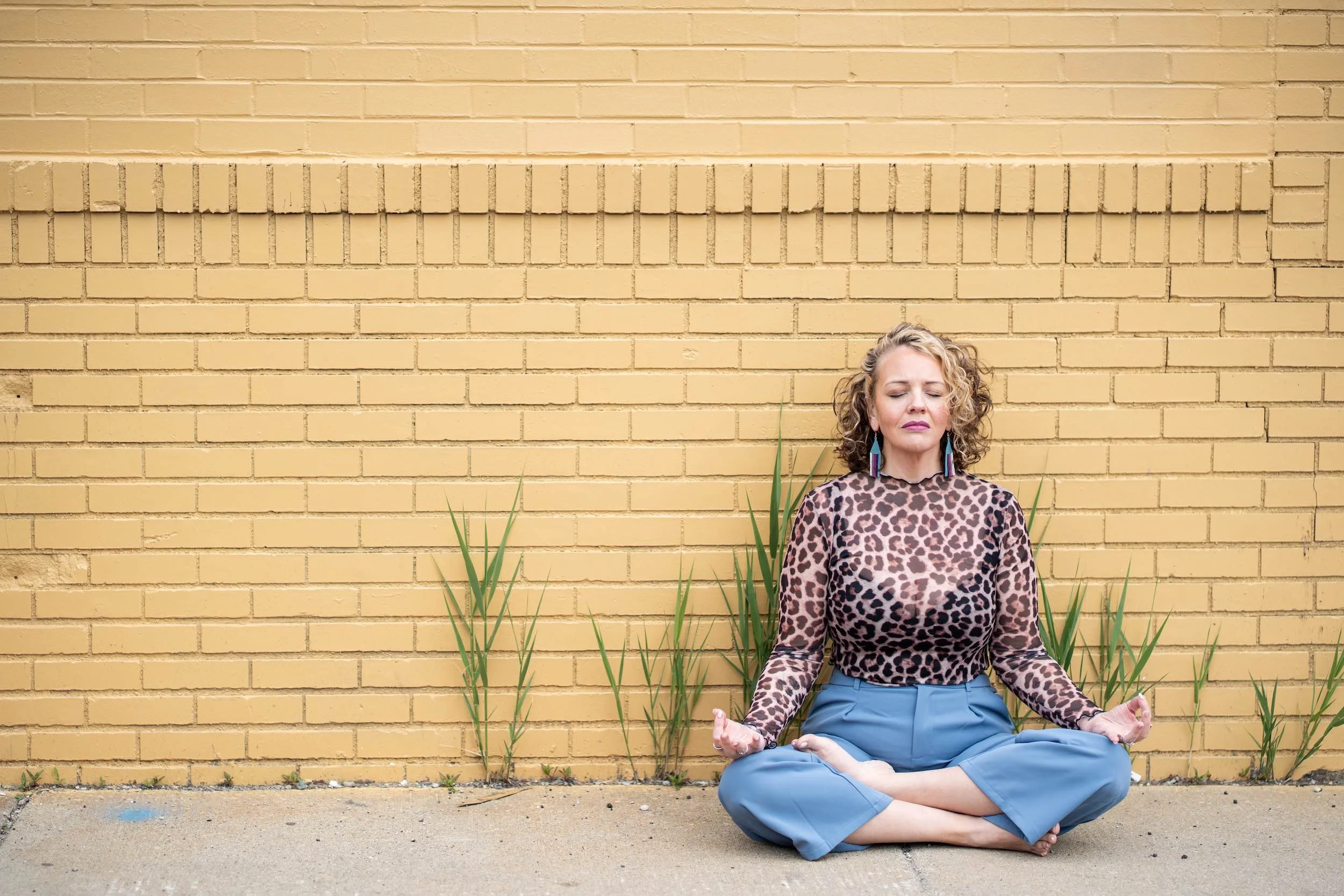 A woman with curly hair in a leopard print top and blue pants sitting cross-legged on the ground in meditation pose against a yellow brick wall with plants surrounding her.