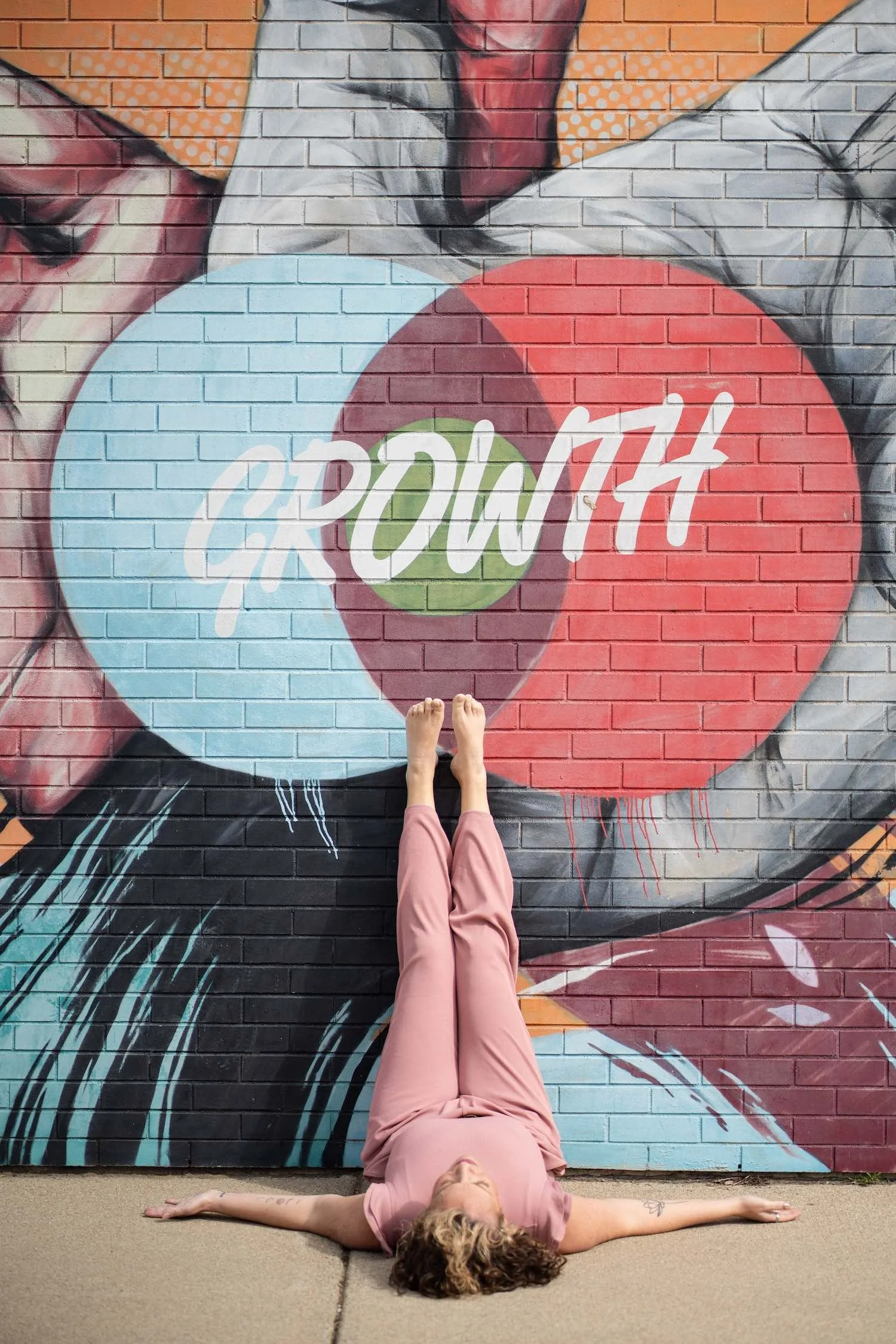 A woman with curly blonde hair lying on the ground in a yoga pose with arms outstretched, on a concrete surface in front of a large colorful mural of a woman's face on a brick wall.