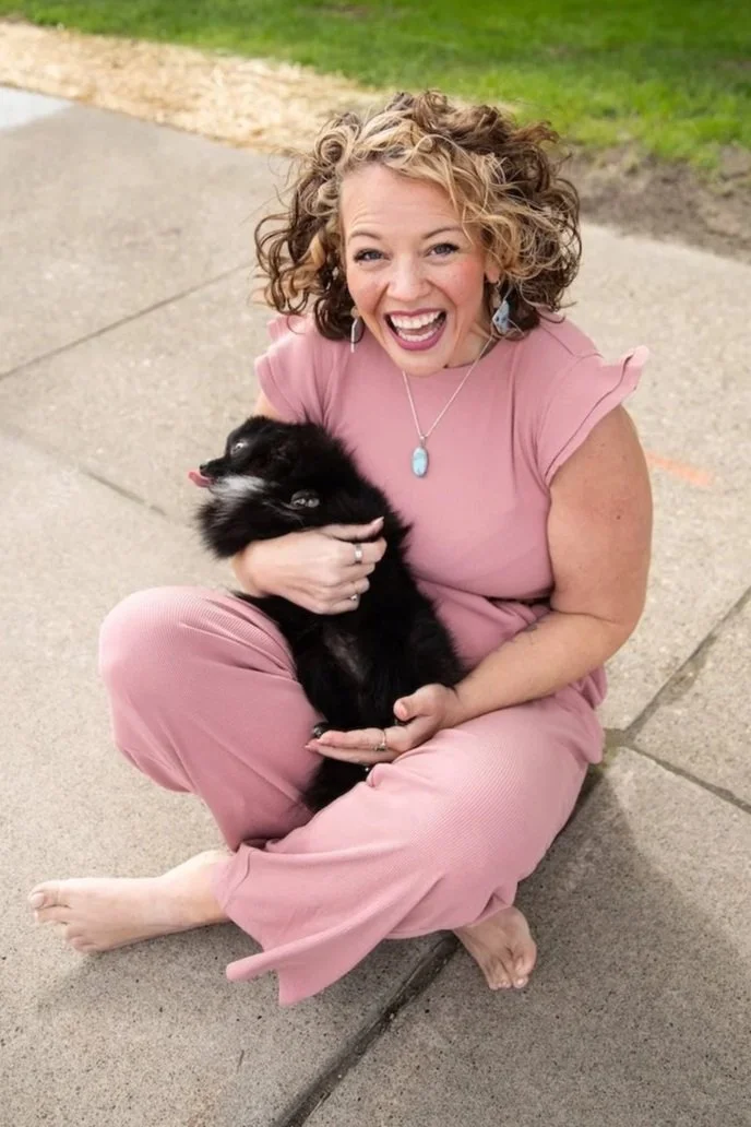 A woman with curly hair, wearing a pink outfit, sitting cross-legged on the sidewalk, holding and playing with a small black dog.