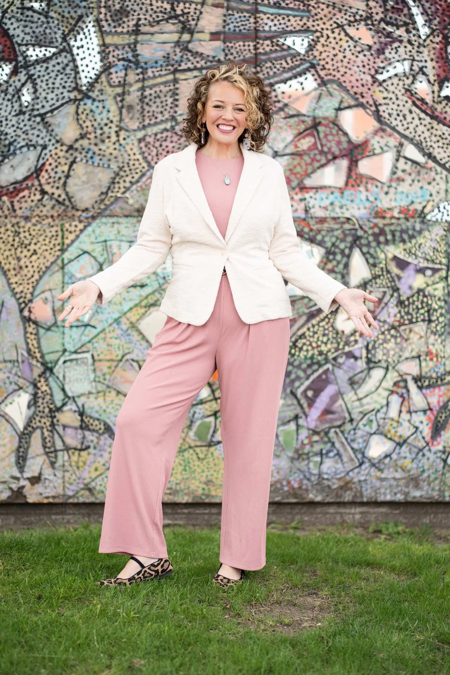 Woman in pink pants, white blazer, pink top, leopard-print shoes, smiling with arms open in front of a colorful mosaic mural.