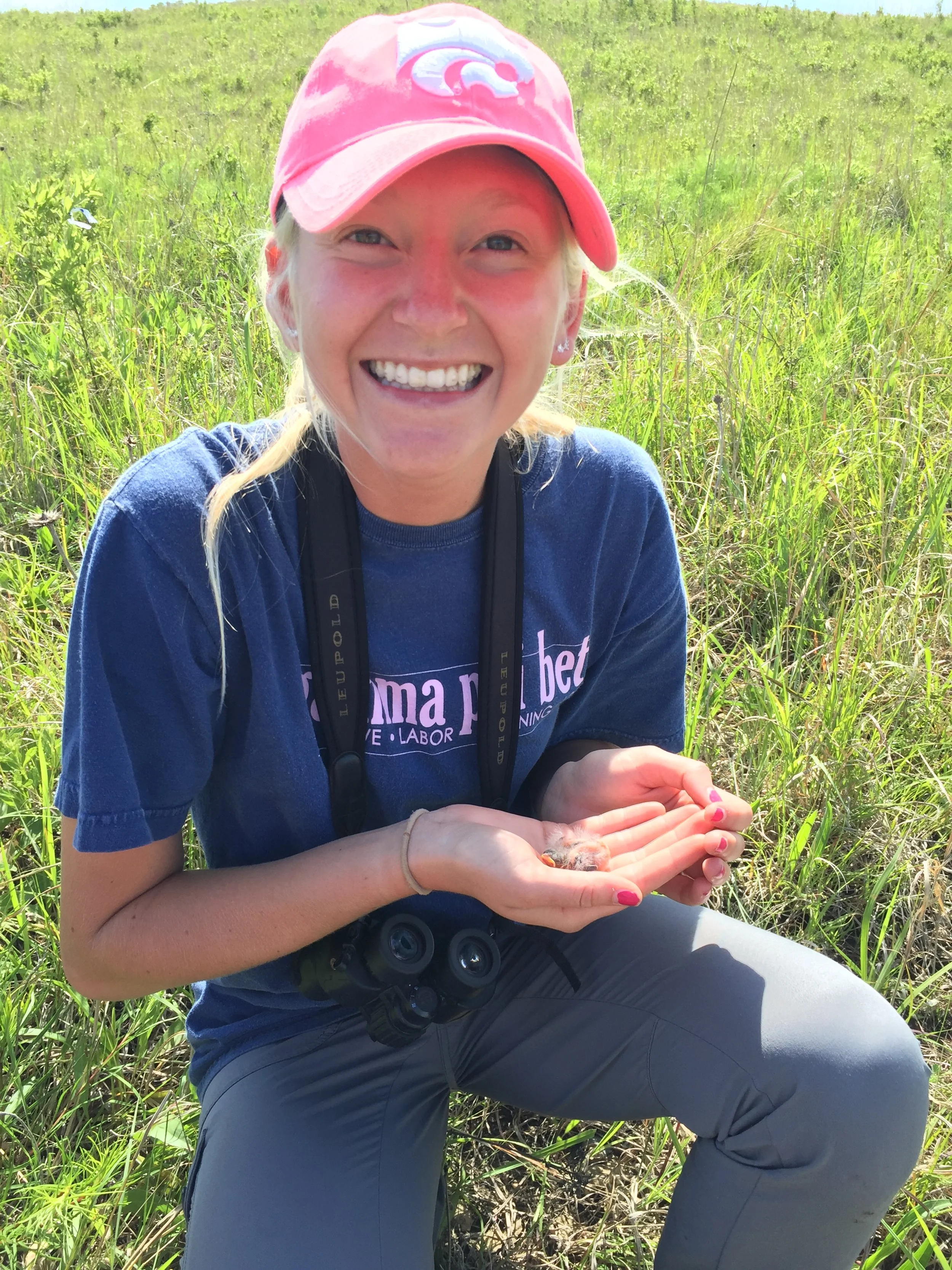 Lauren in a pink cap smiling in a grassy field during her wildlife biology days