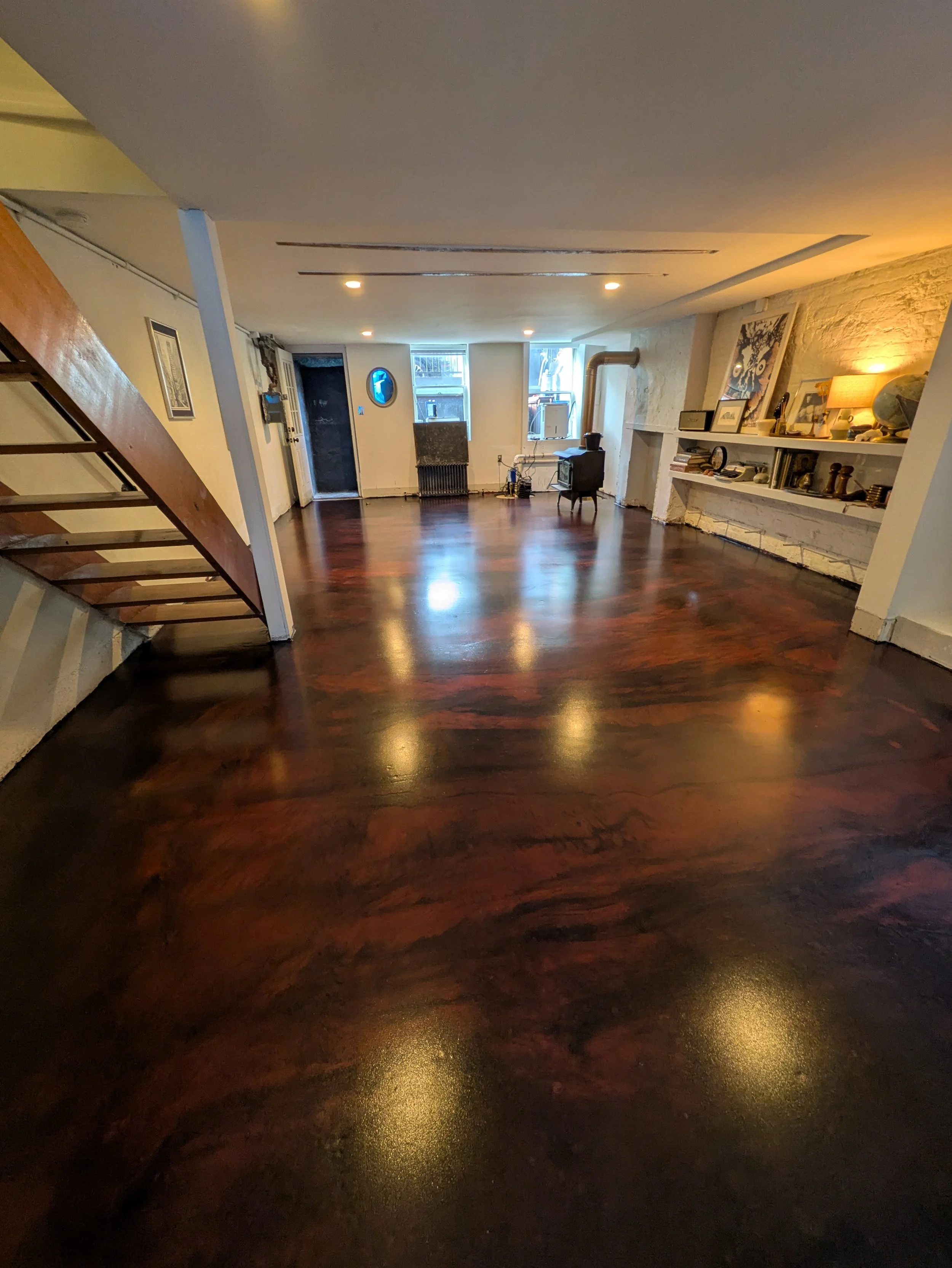 Spacious living room with dark hardwood floors, white brick fireplace, and a stairway on the left. Two windows at the far end allow natural light. Decor includes shelves with various items, framed pictures, and small furnishings.