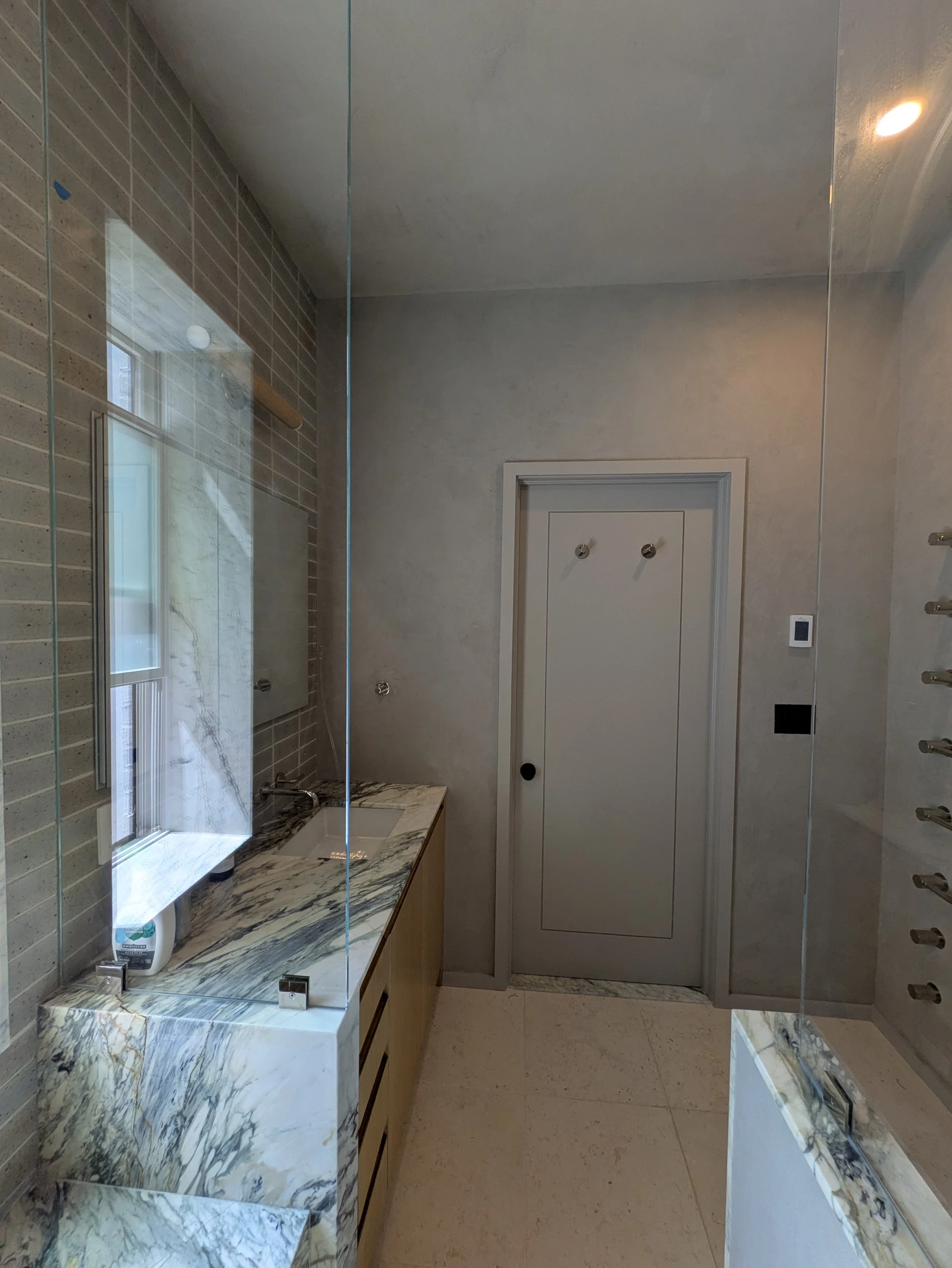 A modern bathroom with a marble countertop, small sink, and a wall-mounted mirror. There is a window with natural light, a closed door, and a wall with built-in shelves or hooks.