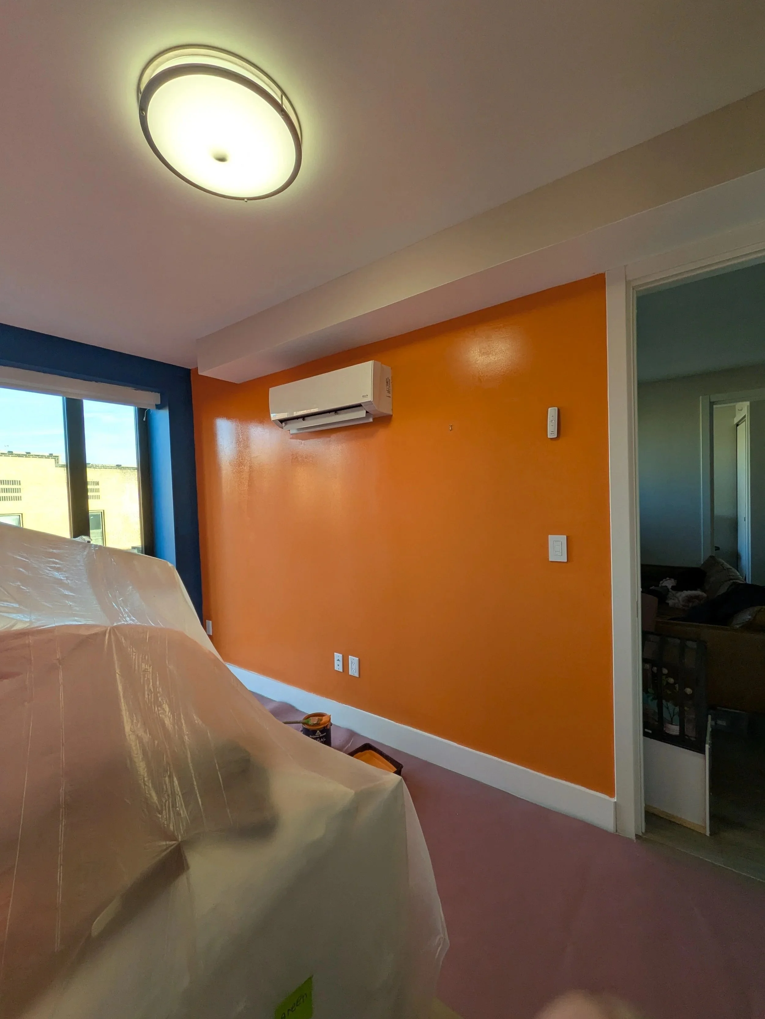 A room with an orange accent wall, a ceiling light, and a window showing a building outside. There is a white air conditioning unit on the wall and a partially covered piece of furniture on the left.