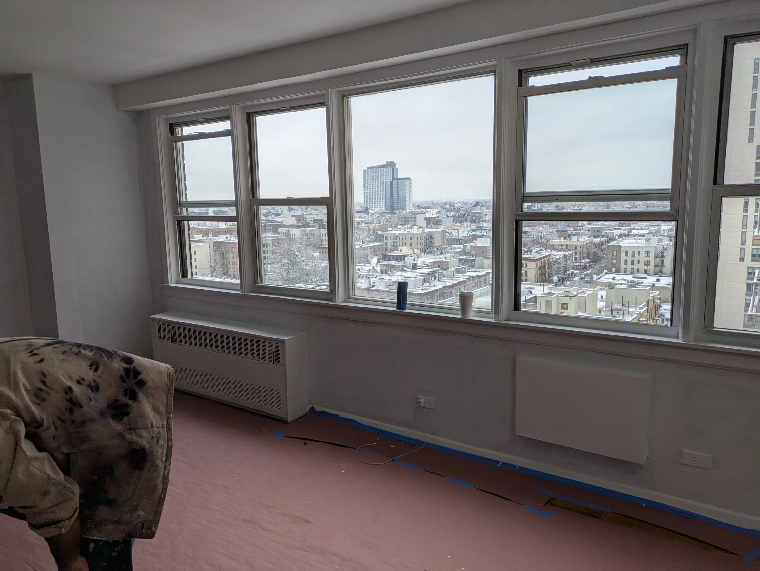Empty living room with large windows showing a snowy cityscape, baseboard heater, and a partially visible couch.