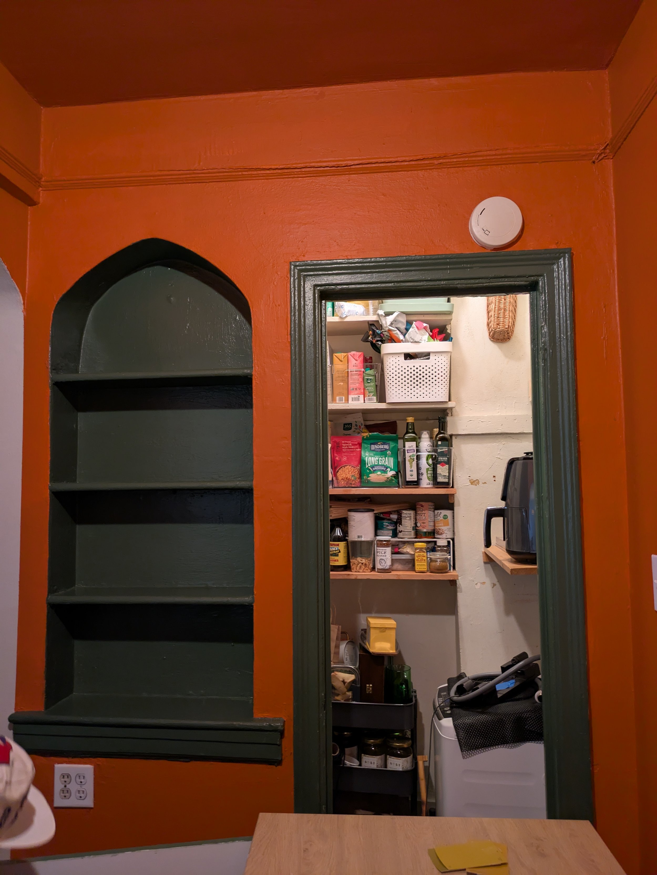 A view of a pantry with shelves filled with food jars, boxes, and bottles, seen through an open door. The surrounding walls are painted orange with green trim around the door.