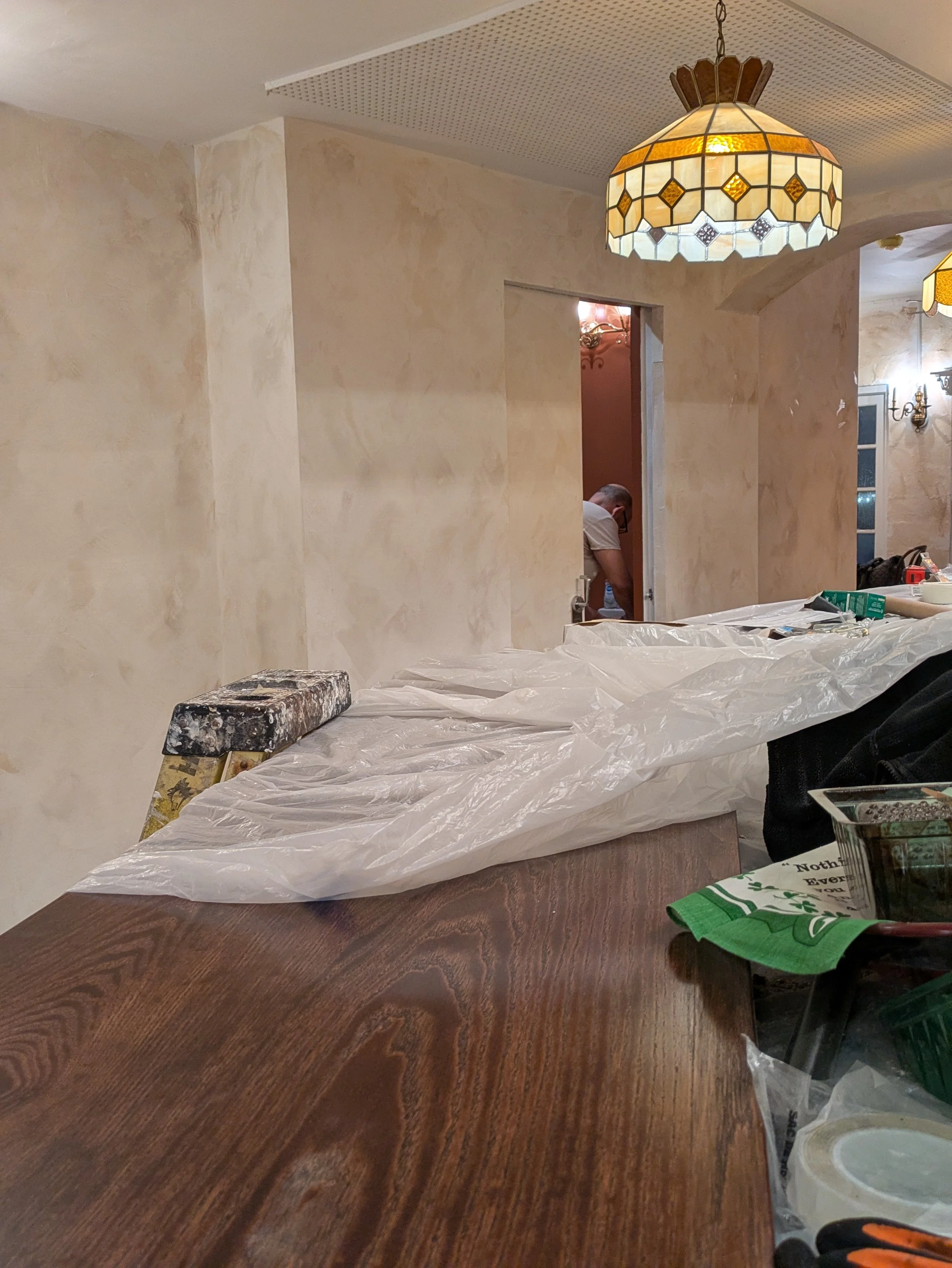 Interior of a room undergoing renovation with unfinished walls, hanging pendant lights, and a table covered with plastic and construction supplies. A worker is visible in the background.