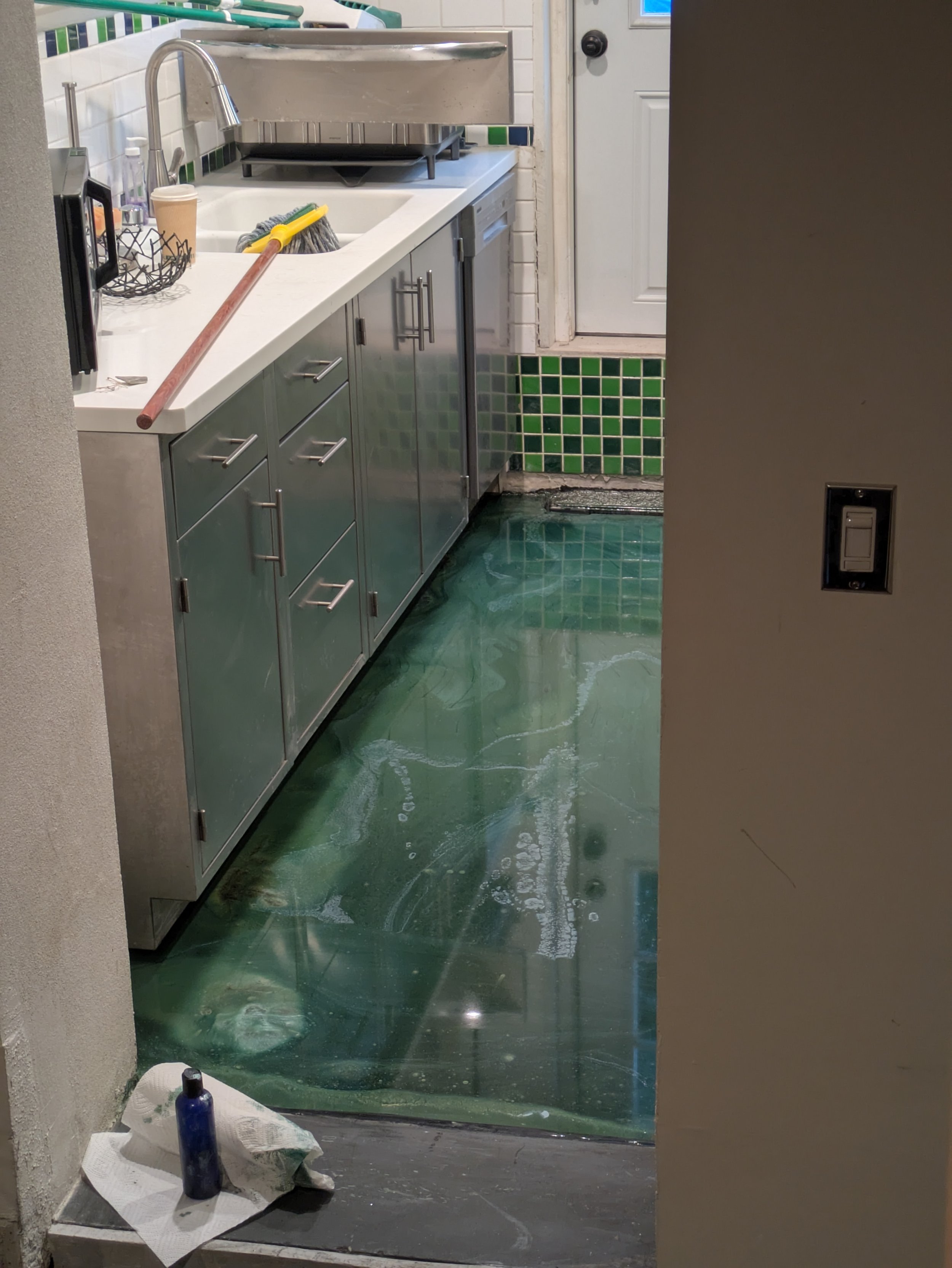 Kitchen floor with green epoxy coating and a small section of gray concrete at the entrance. The countertop holds cleaning supplies, including a mop with a yellow handle, a spray bottle, and paper towels. The sink area has a stainless steel sink, a b