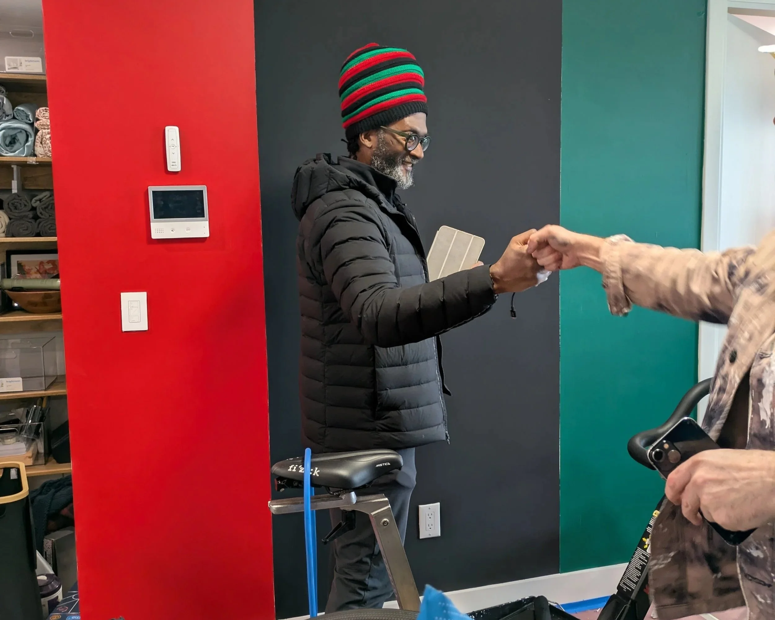 A man wearing glasses, a colorful striped beanie, and a black puffy jacket is fist-bumping a woman who is holding a phone, in an indoor setting with colorful walls and shelves with supplies in the background.
