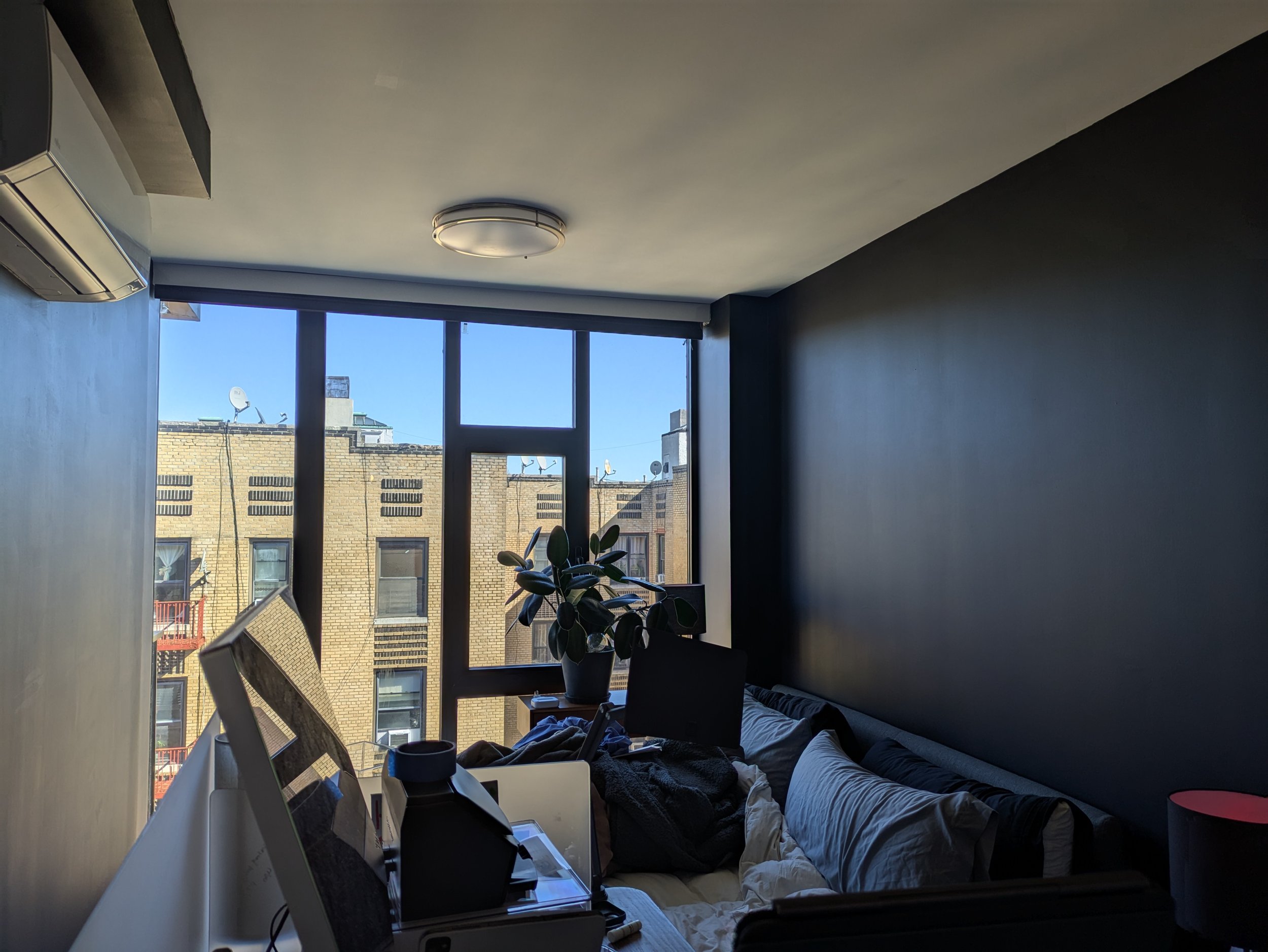 A messy bedroom with a computer desk, a plant, a bed, and large windows showing neighboring buildings.