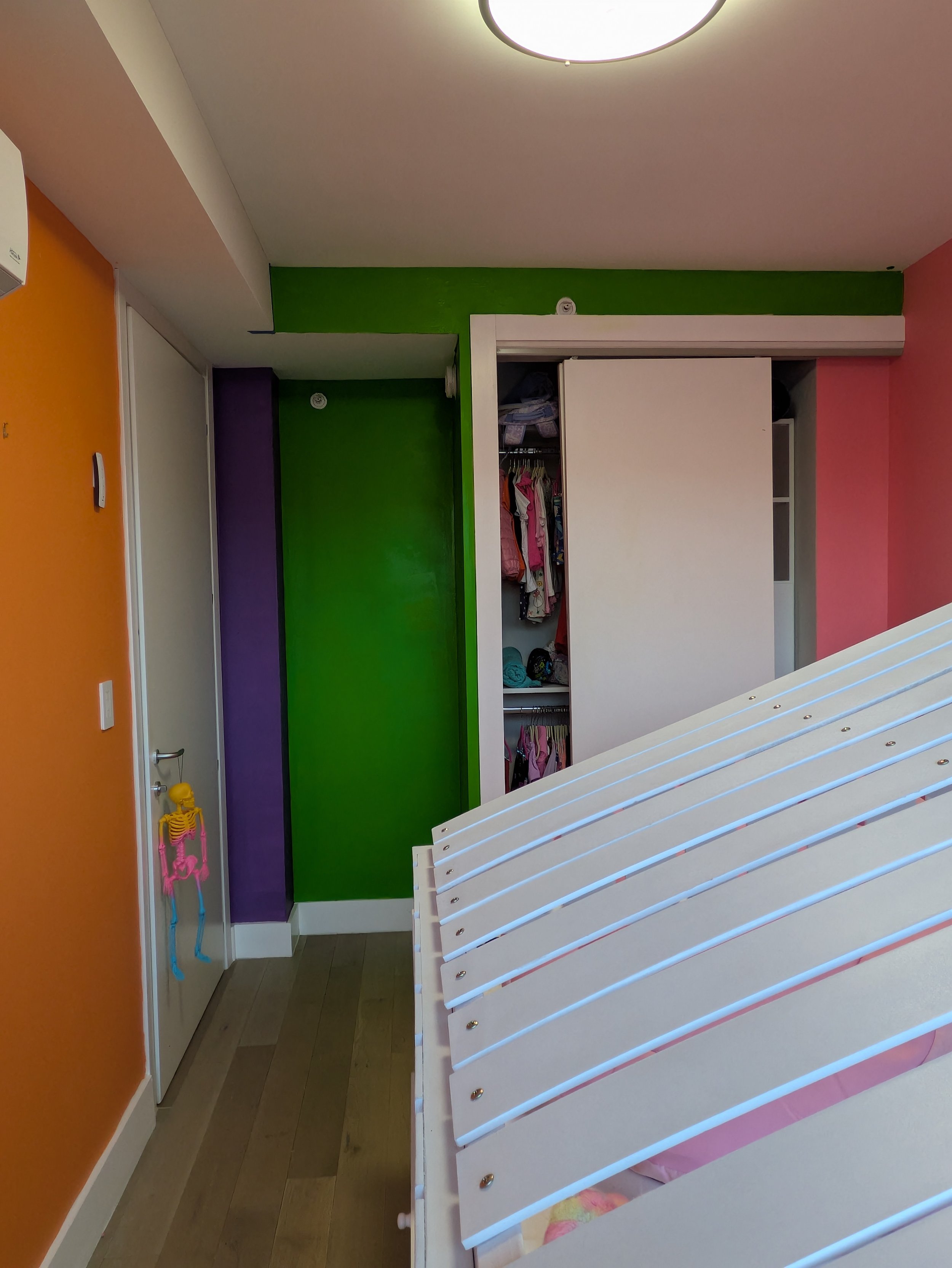 Colorful bedroom with orange, purple, green, and pink walls. An open closet with clothes inside, next to a slatted bed frame without a mattress. The room has a ceiling light and a skeleton decoration hanging on the door handle.