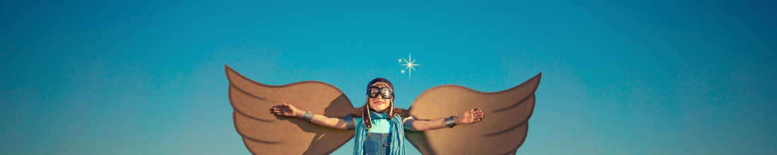 Child dressed as a superhero with cardboard wings and goggles, standing against a bright blue sky.