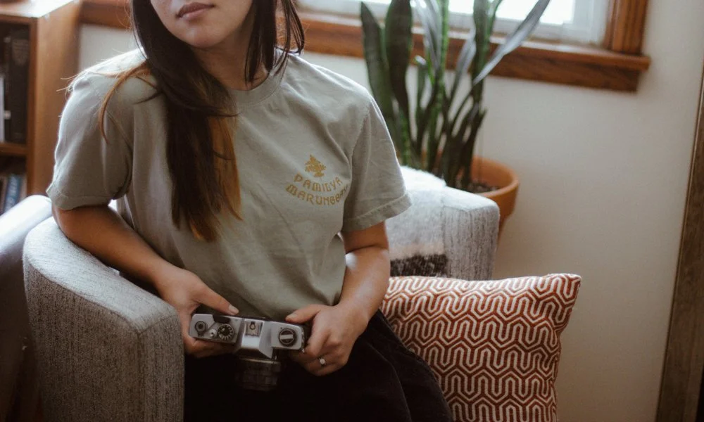 A young woman sitting on a beige armchair holding a camera in her lap, with a patterned pillow and a potted plant in the background.