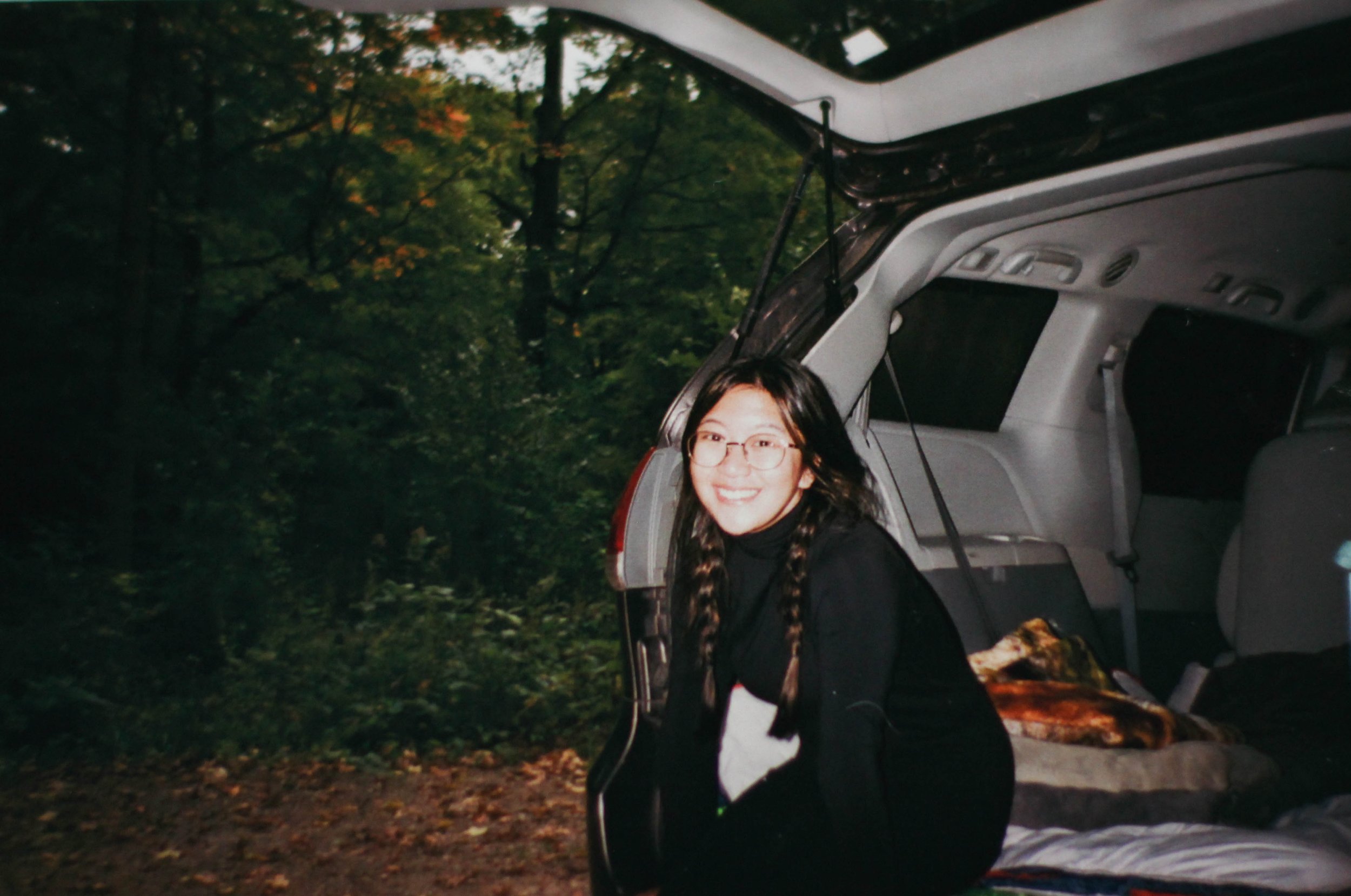 A young woman with glasses and braided hair smiling as she leans out of the open trunk of a vehicle, outdoors with trees in the background.