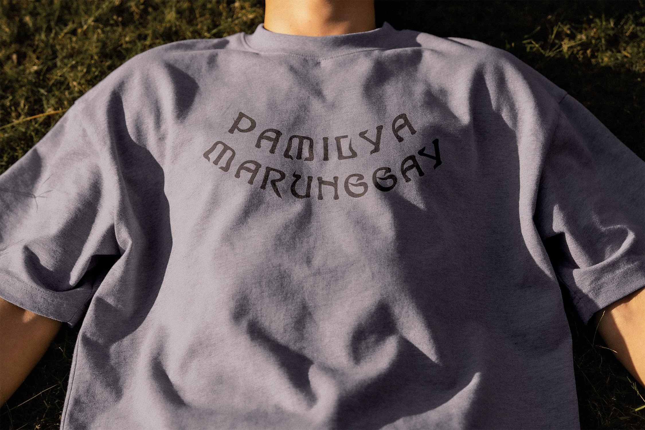 A person lying on the grass wearing a gray T-shirt with the words 'PAMILYA' and 'MARUNGGAY' printed on the front.