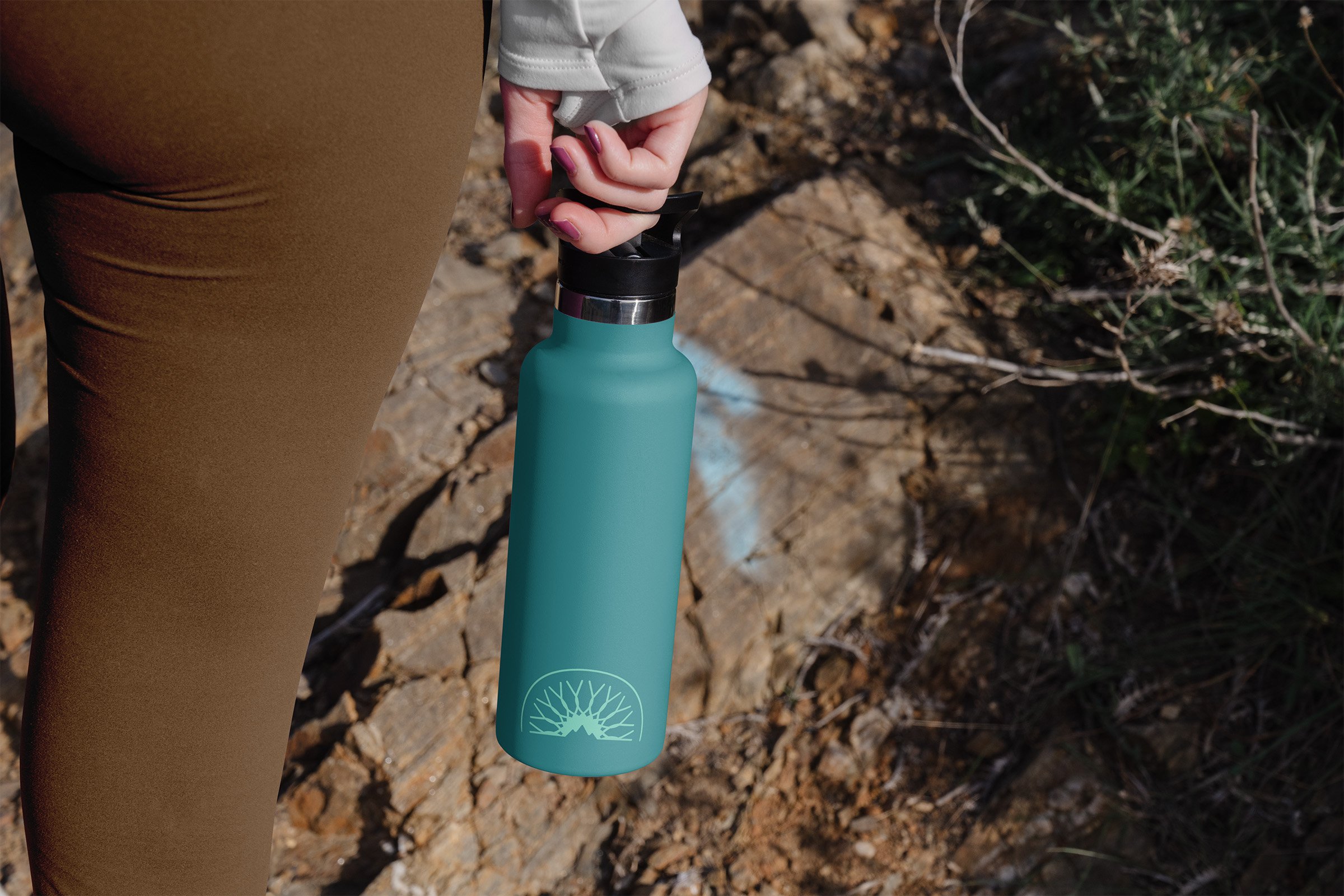 A person holding a turquoise water bottle with a black lid, standing outdoors on rocky ground with some green vegetation in the background.