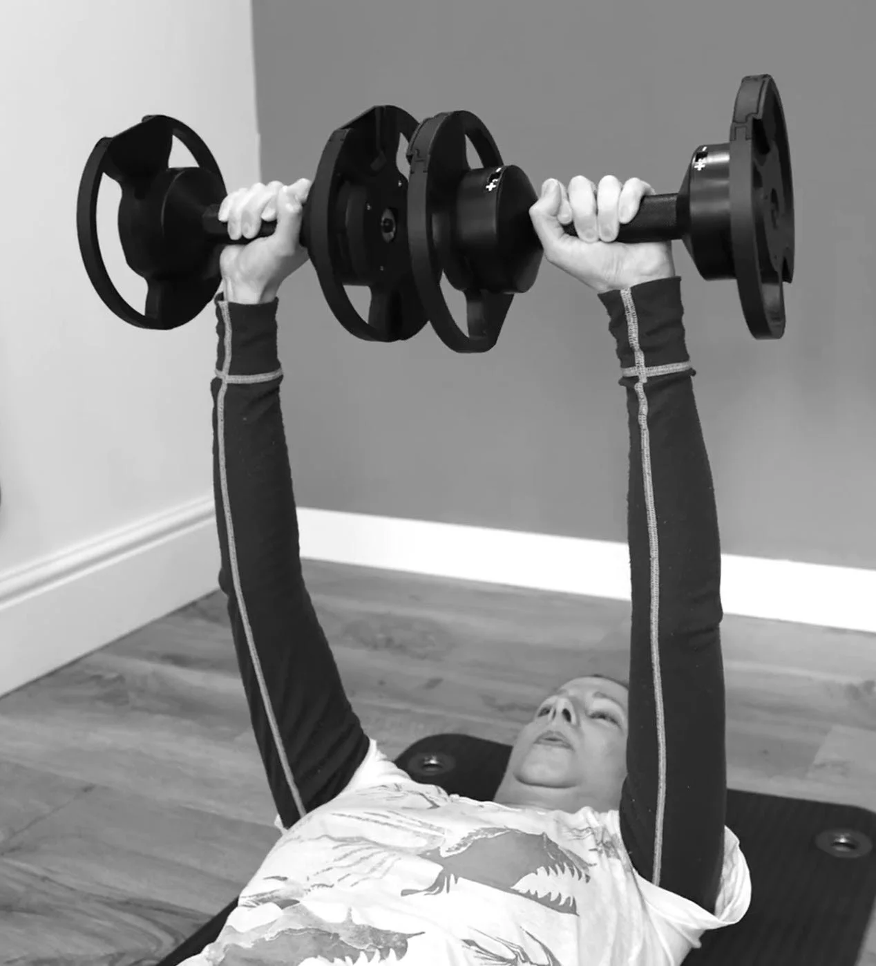 A person lying on a gym mat lifting a barbell with weights, wearing long-sleeve athletic shirt.