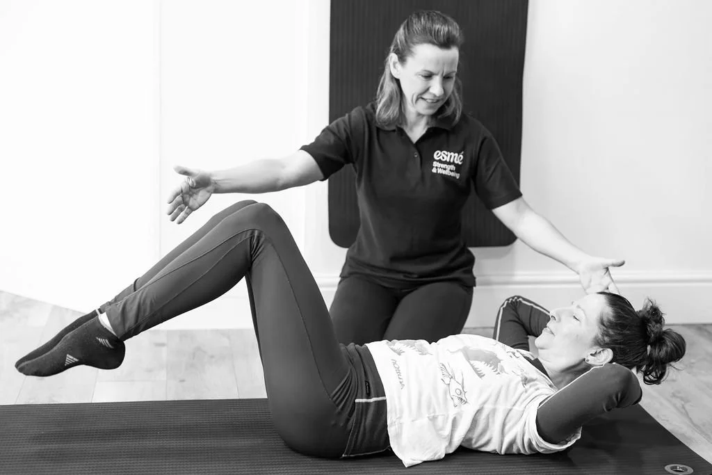 A woman is lying on her back on a yoga mat, doing a core exercise with her knees bent, while another woman, likely an instructor, assists or guides her in a fitness or physical therapy session.
