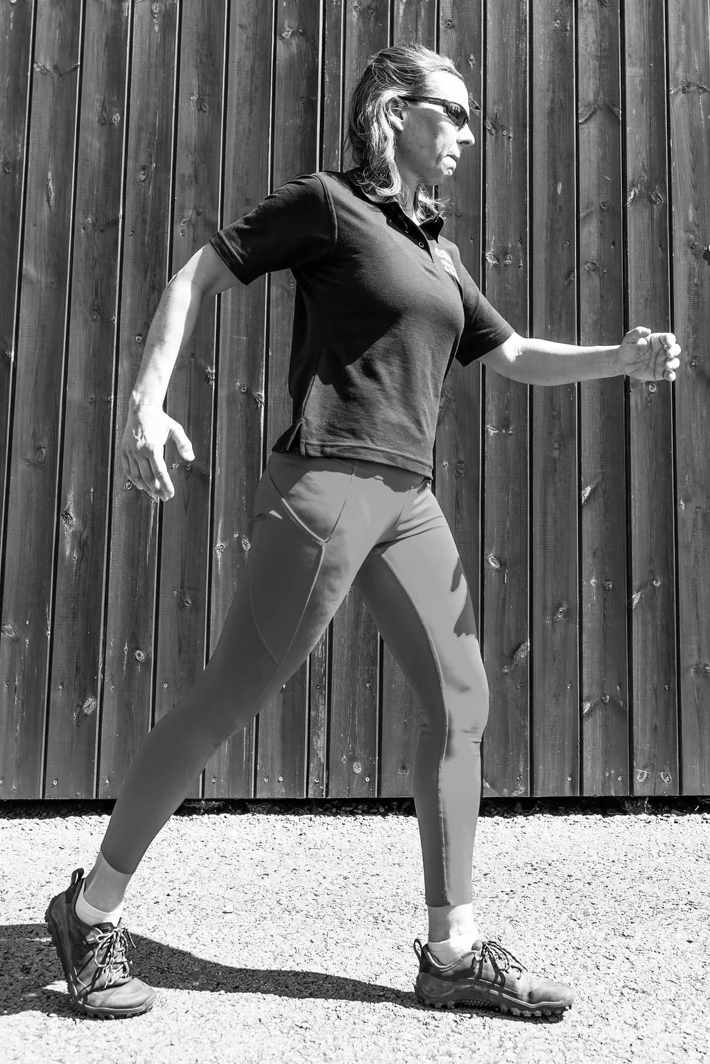 A woman outdoors, wearing athletic gear, walking on a gravel surface in front of a wooden fence, with her hair tied back and sunglasses on.