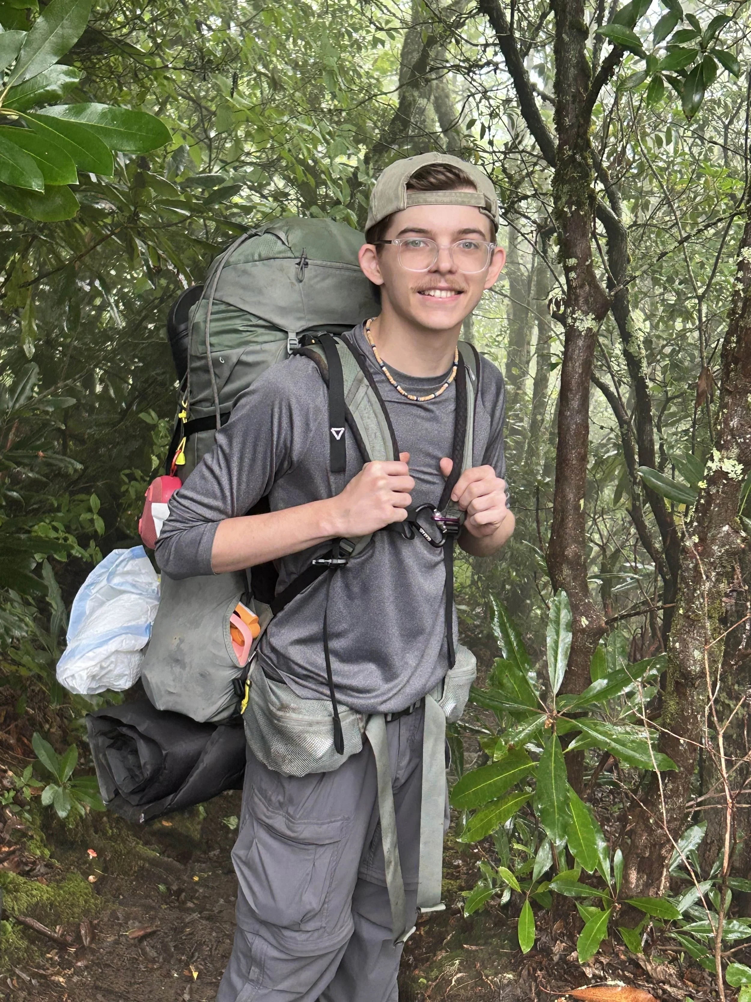 Person with glasses and a cap hiking in a lush green forest, carrying a large backpack and wearing outdoor gear.