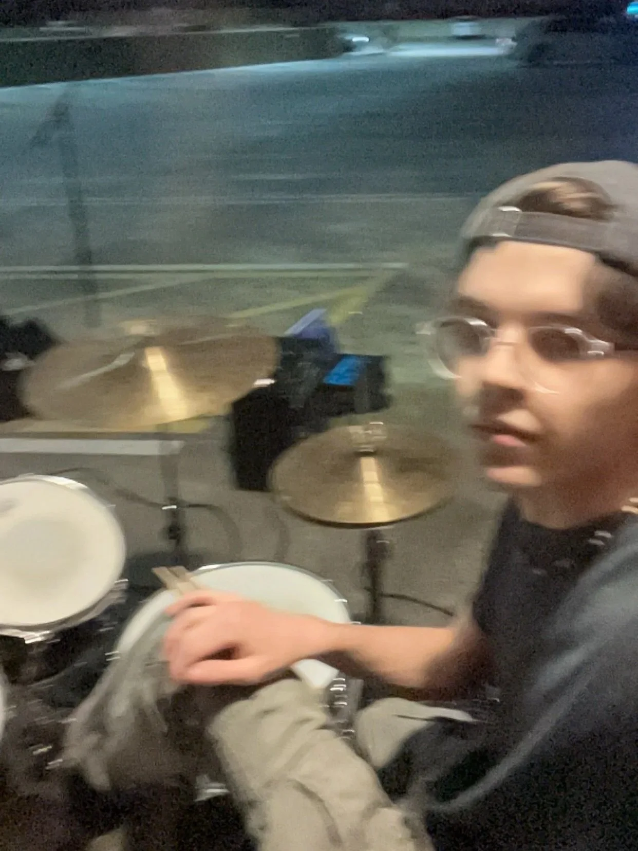 A young man wearing glasses and a backward cap sitting at a drum set, holding drumsticks, inside a vehicle at night.