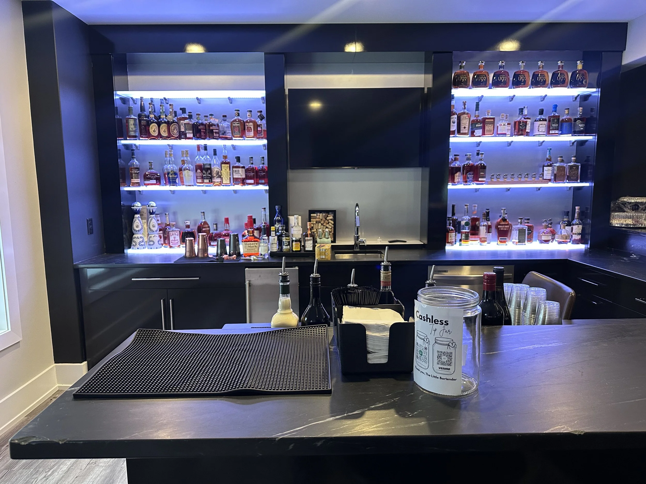 A bar counter with a black countertop holding glasses, bottles of cocktail ingredients, and signs, with a backlit display of various liquor bottles on shelves.