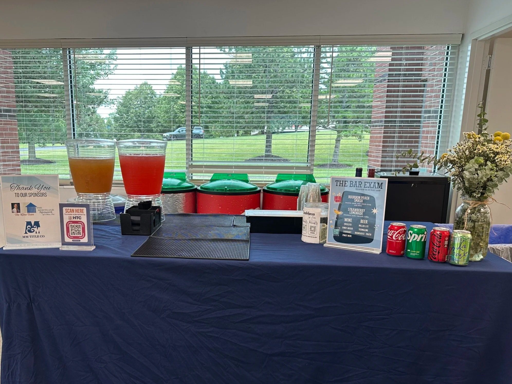 Ice and fruit drinks on a table in front of a large window with trees outside. The drinks are in clear glasses, one yellowish and one red. There are red containers, promotional signs, soda cans, a small loose pile of clear plastic cups, and a floral arrangement on the table.