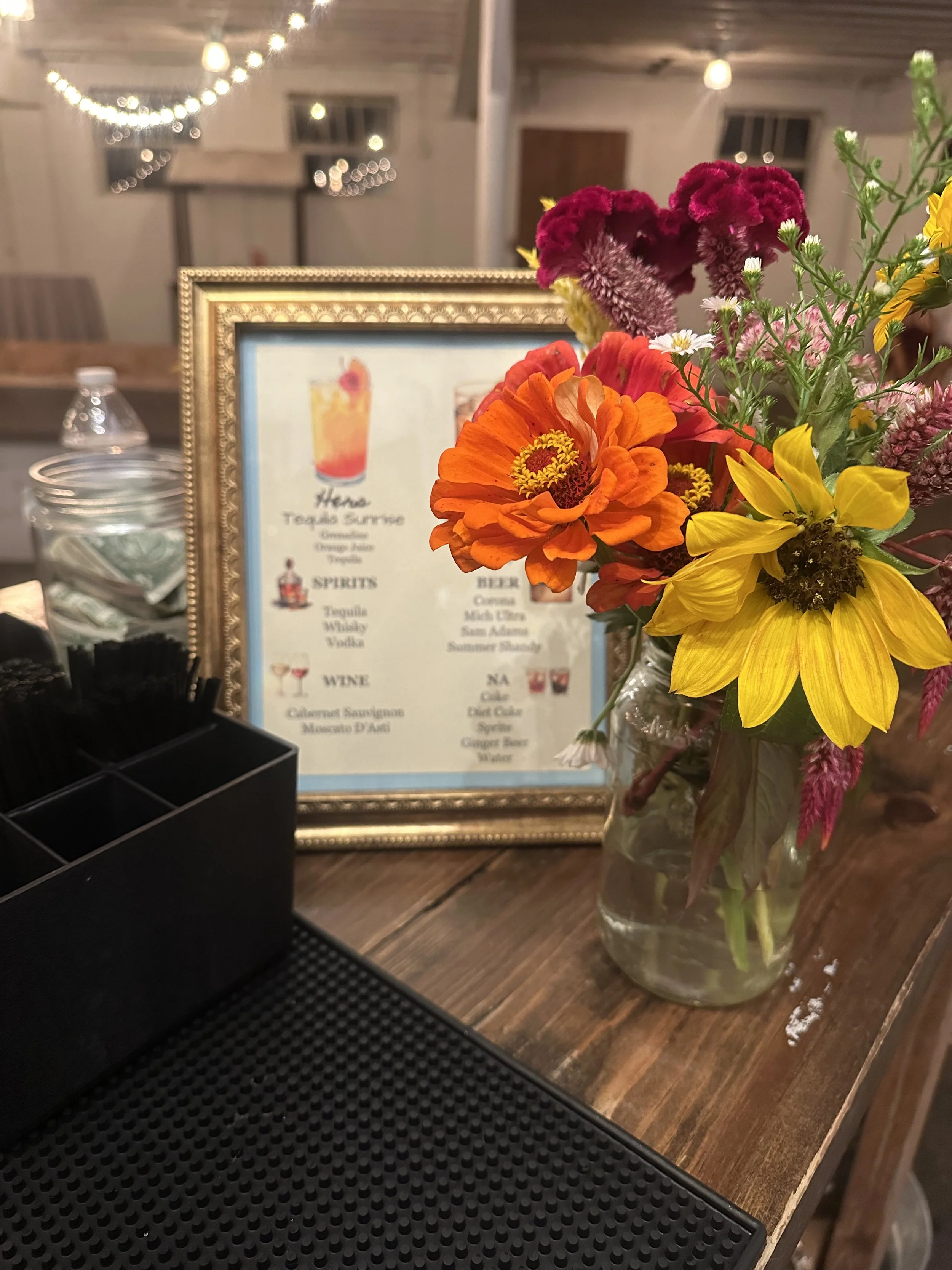 A glass jar filled with colorful flowers including orange, yellow, purple, and white blooms on a wooden surface, with a gold framed menu in the background and string lights hanging overhead.