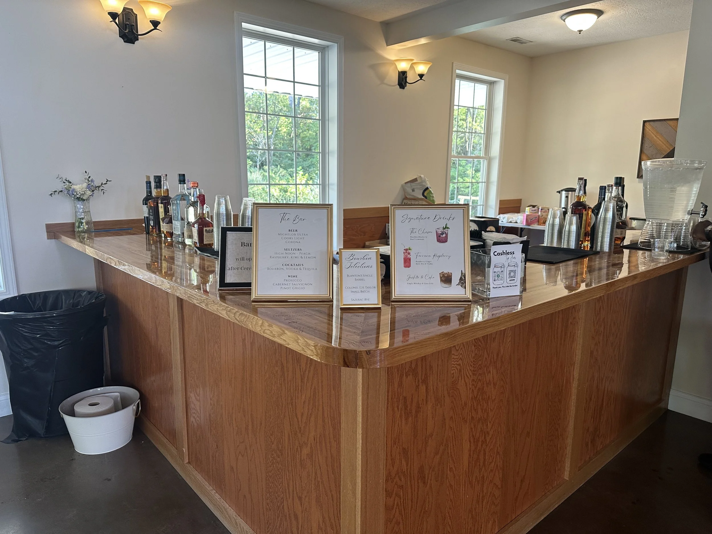A wooden bar counter with various bottles of alcohol, drinks menu, and beverage containers inside a room with large windows and ceiling lights.