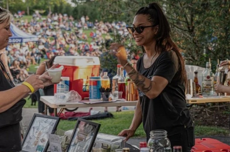 A woman with sunglasses and tattoos serving drinks and bartending at an outdoor event, with a crowd sitting on a grassy hill in the background.