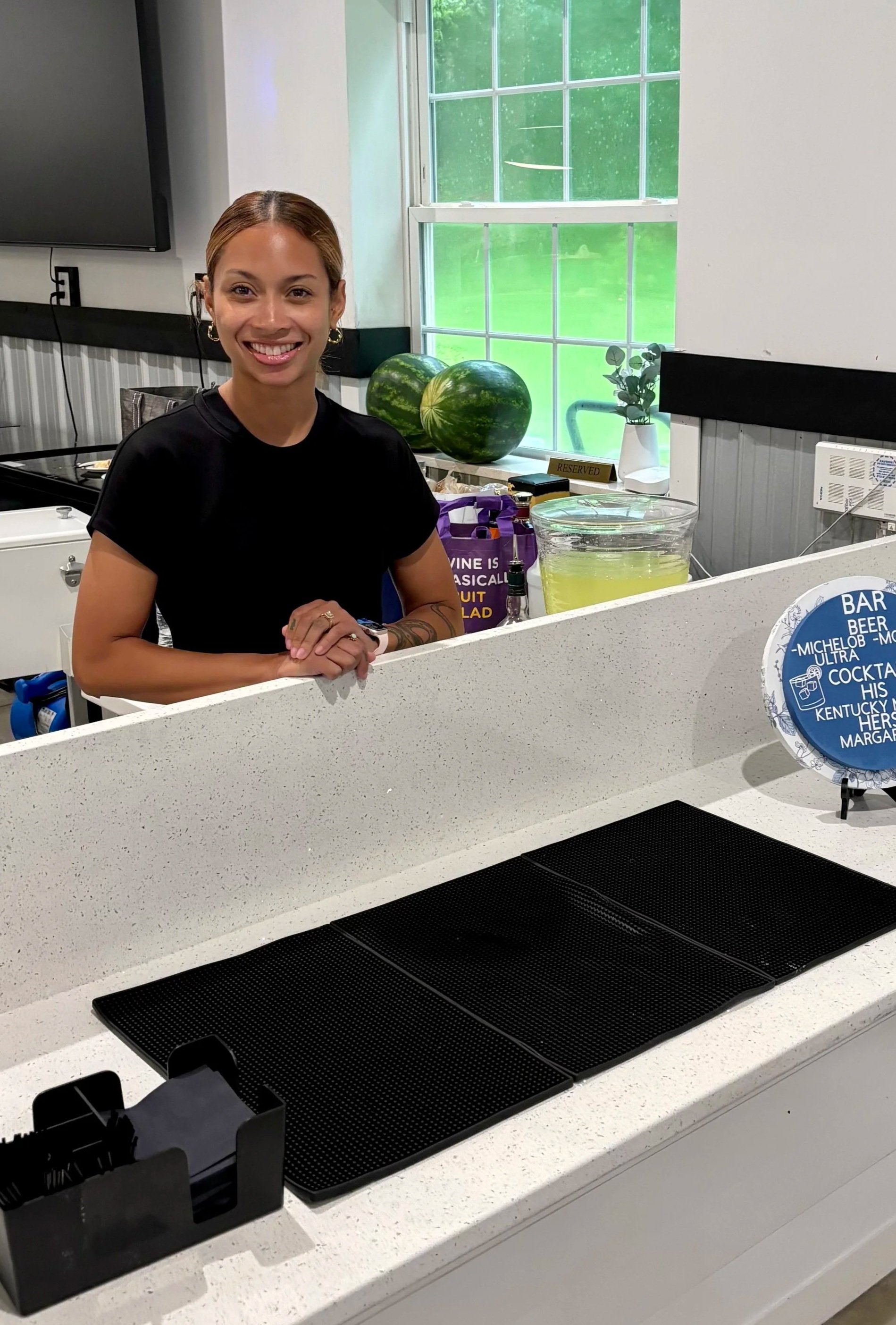 A woman with brown hair tied back, wearing a black shirt and jewelry, standing behind a white countertop in a room with large windows, watermelons, a potted plant, and a reserved sign on the counter. bartender