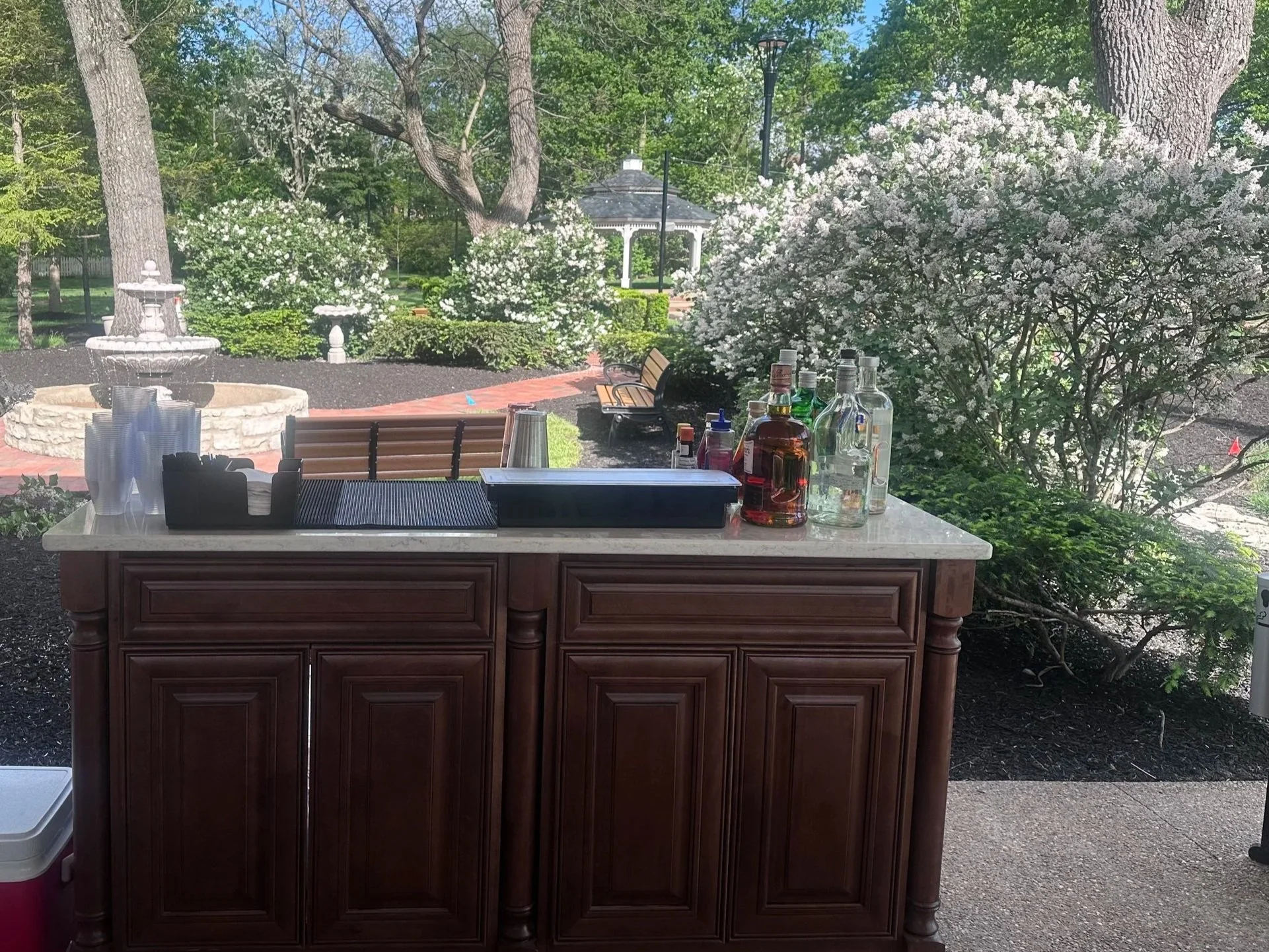 Outdoor scene with a bar table in the foreground, bottles of alcohol, cups, and napkins. In the background, a garden with trees, bushes, a fountain, and a gazebo.