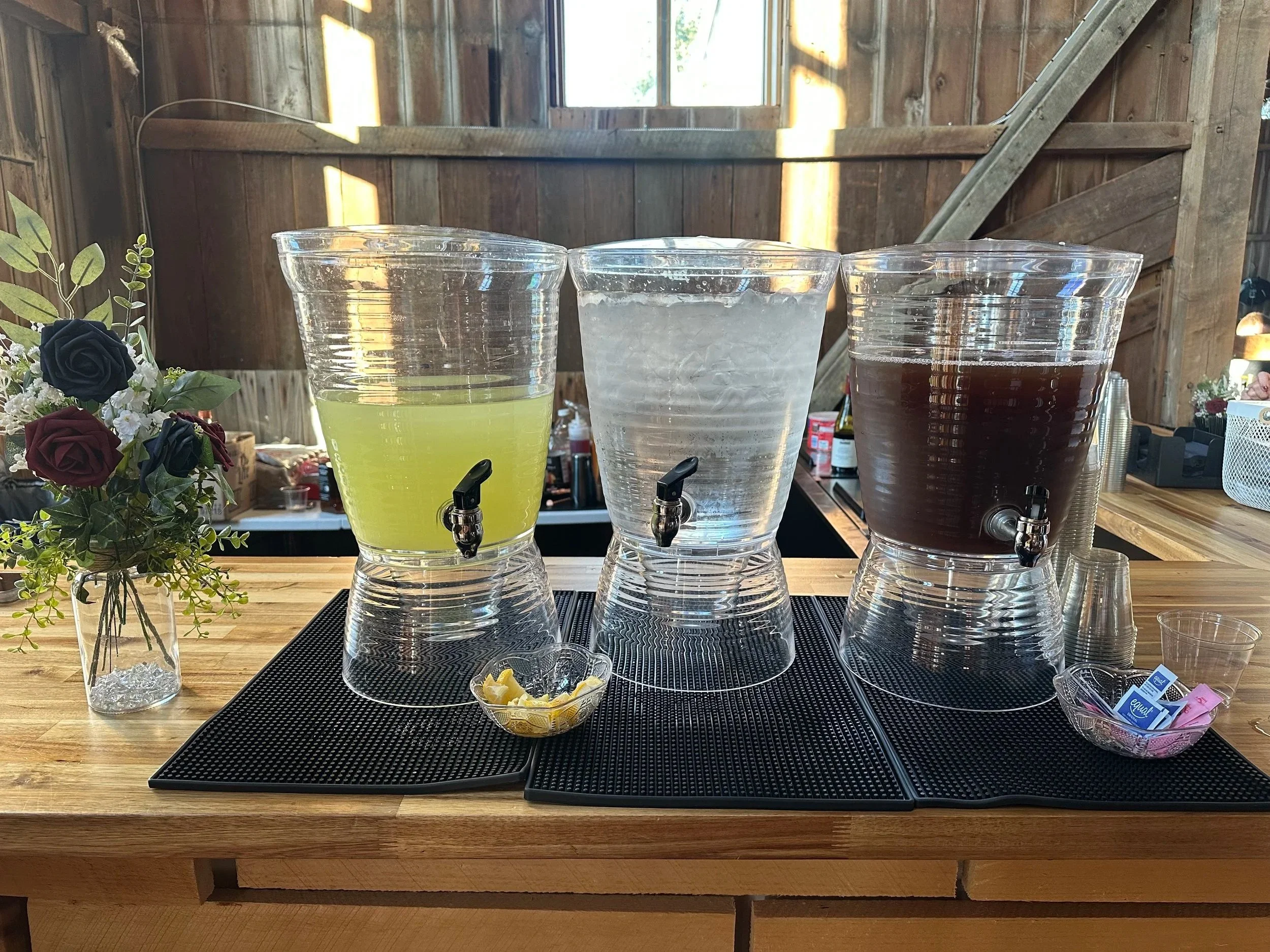 Three large glass beverage dispensers on a wooden counter, filled with colorful drinks. The left dispenser contains a light yellow beverage, the middle one appears to be empty or frosted, and the right one has a dark brown drink. There is a small bowl of lemon slices in front of the dispensers. To the left, there is a small vase with a bouquet of dark red, black, and white roses with greenery. In the background, the setting appears to be a rustic indoor space with wooden walls, a window, and some decorative items.